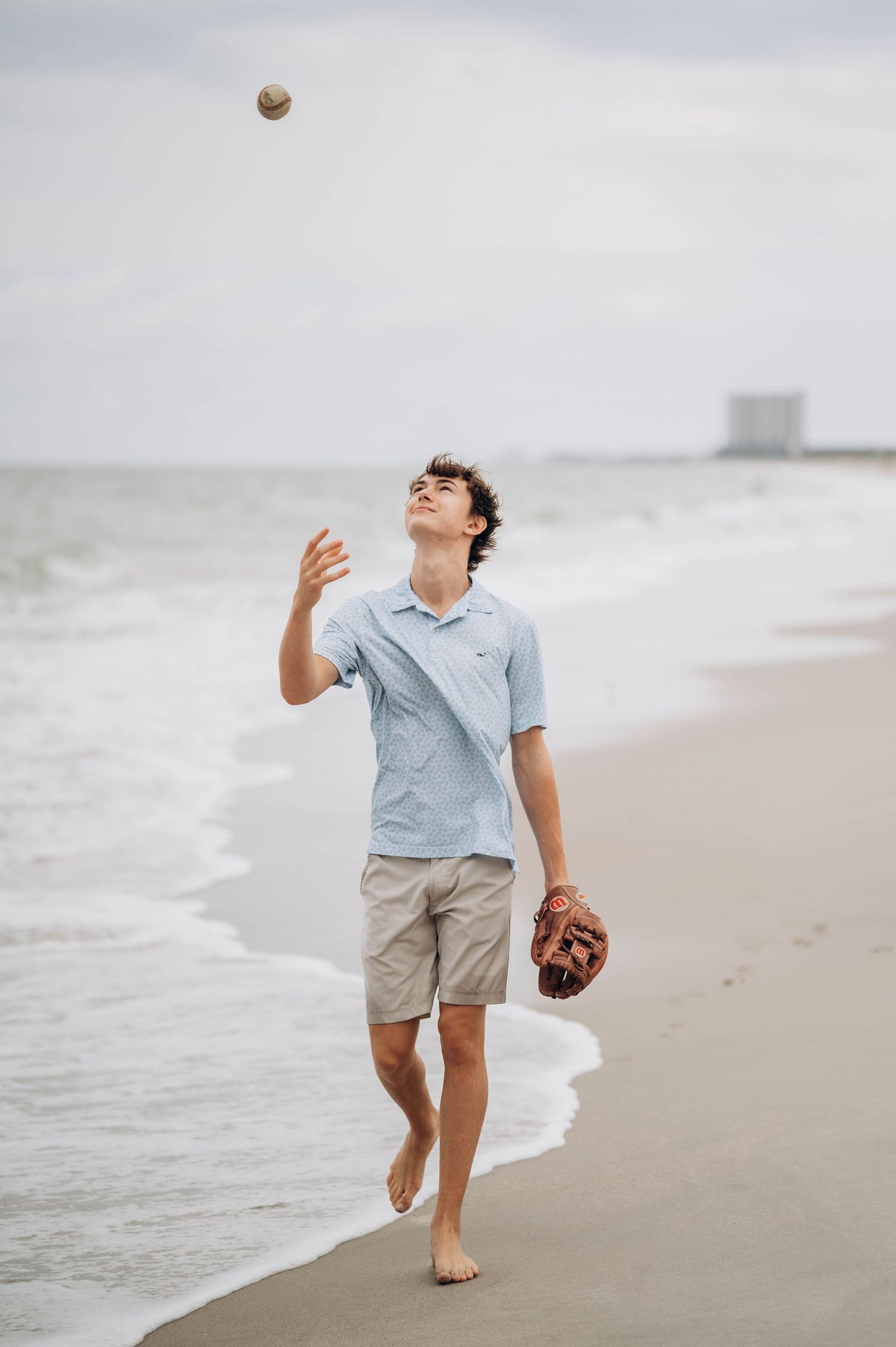 Person playing baseball alone on a cloudy beach, tossing and catching the ball.