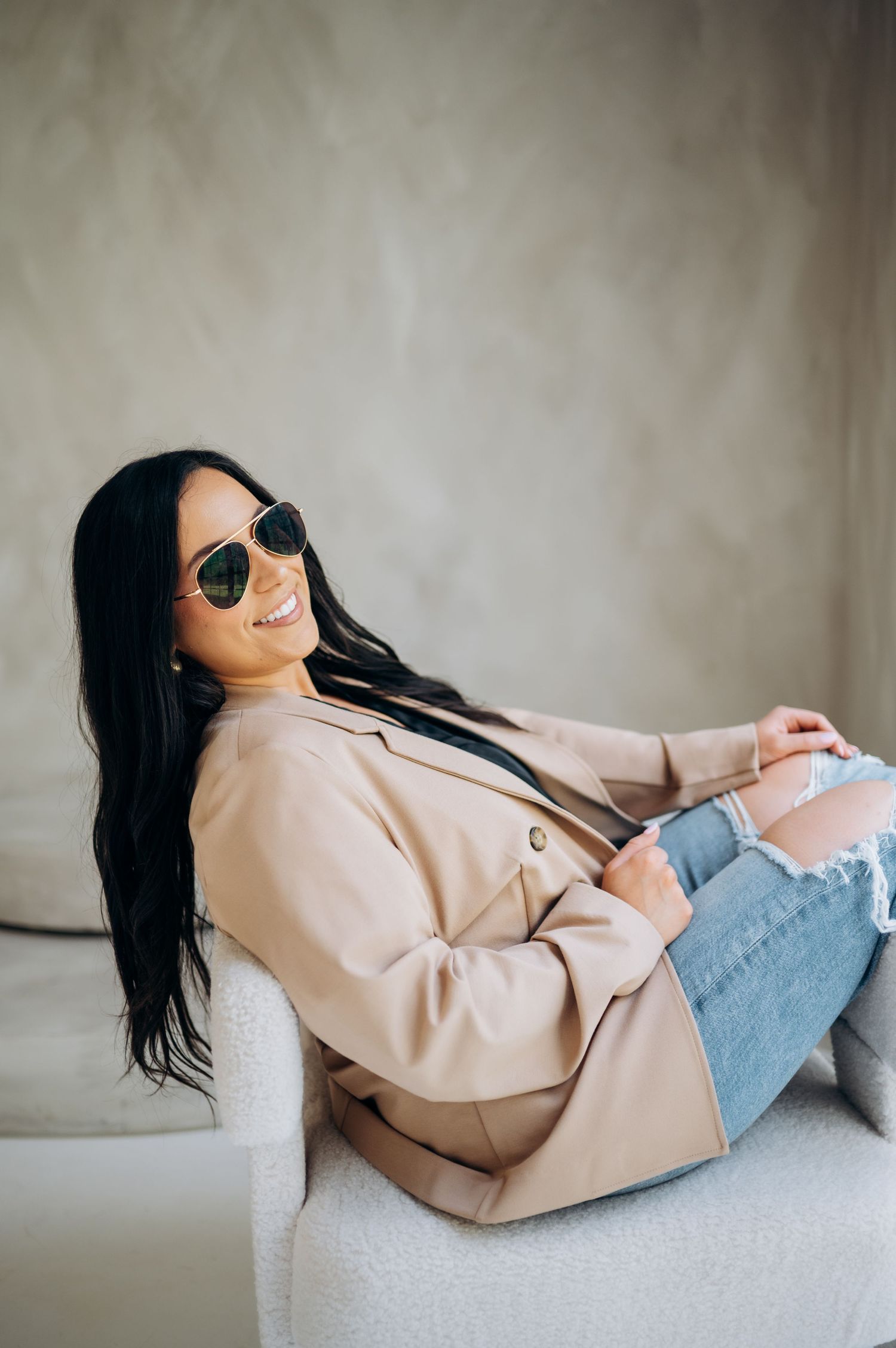 Casual chic look with beige blazer and distressed denim while relaxing on white chair.