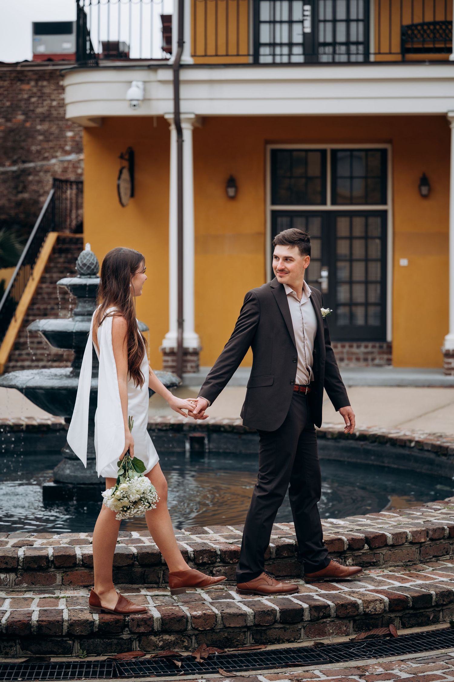 Newlywed couple holding hands and walking along the edge of a brick fountain outside a yellow building.