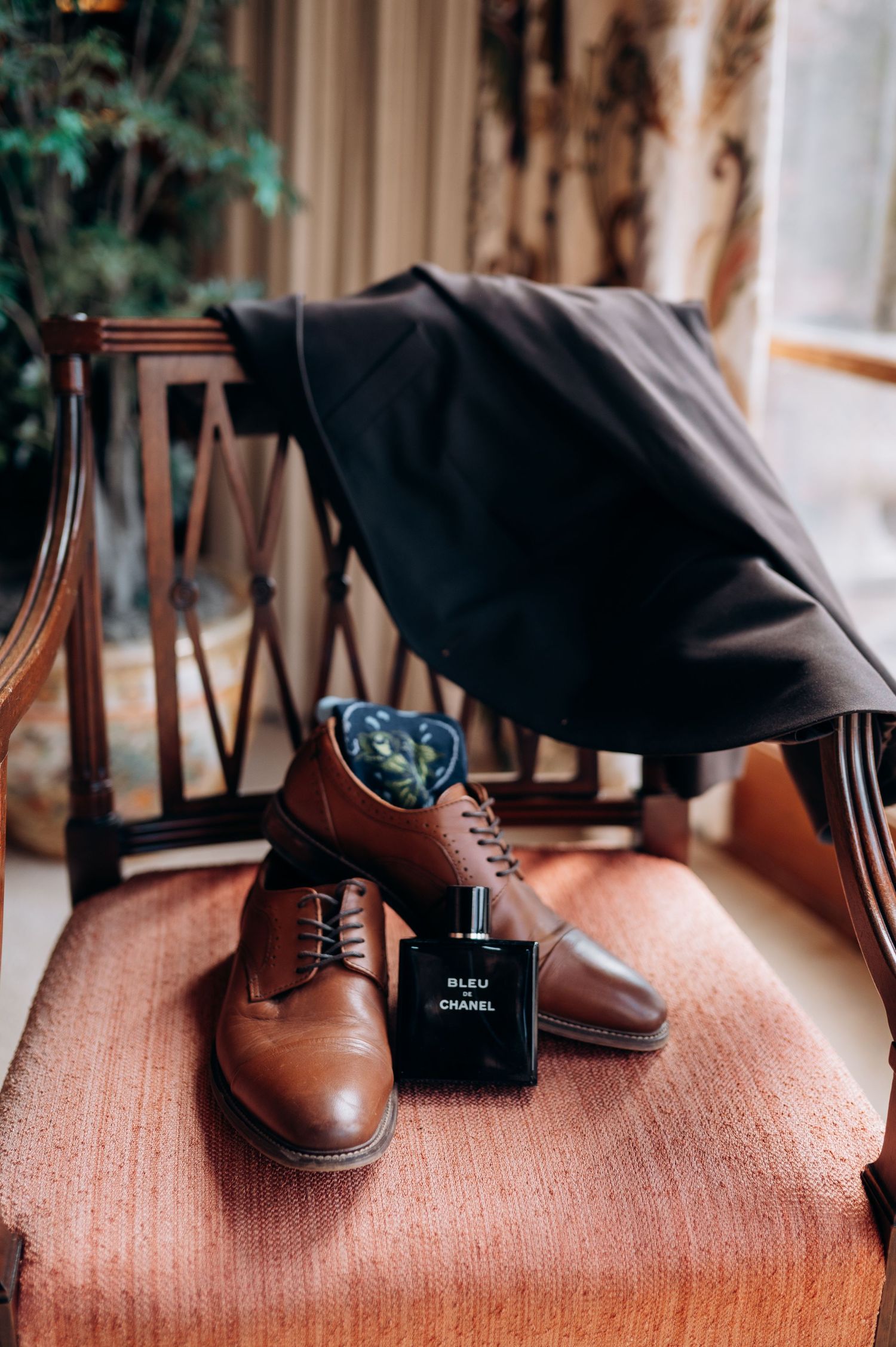 Brown leather Oxford shoes with Bleu de Chanel cologne and dark suit on a vintage armchair.