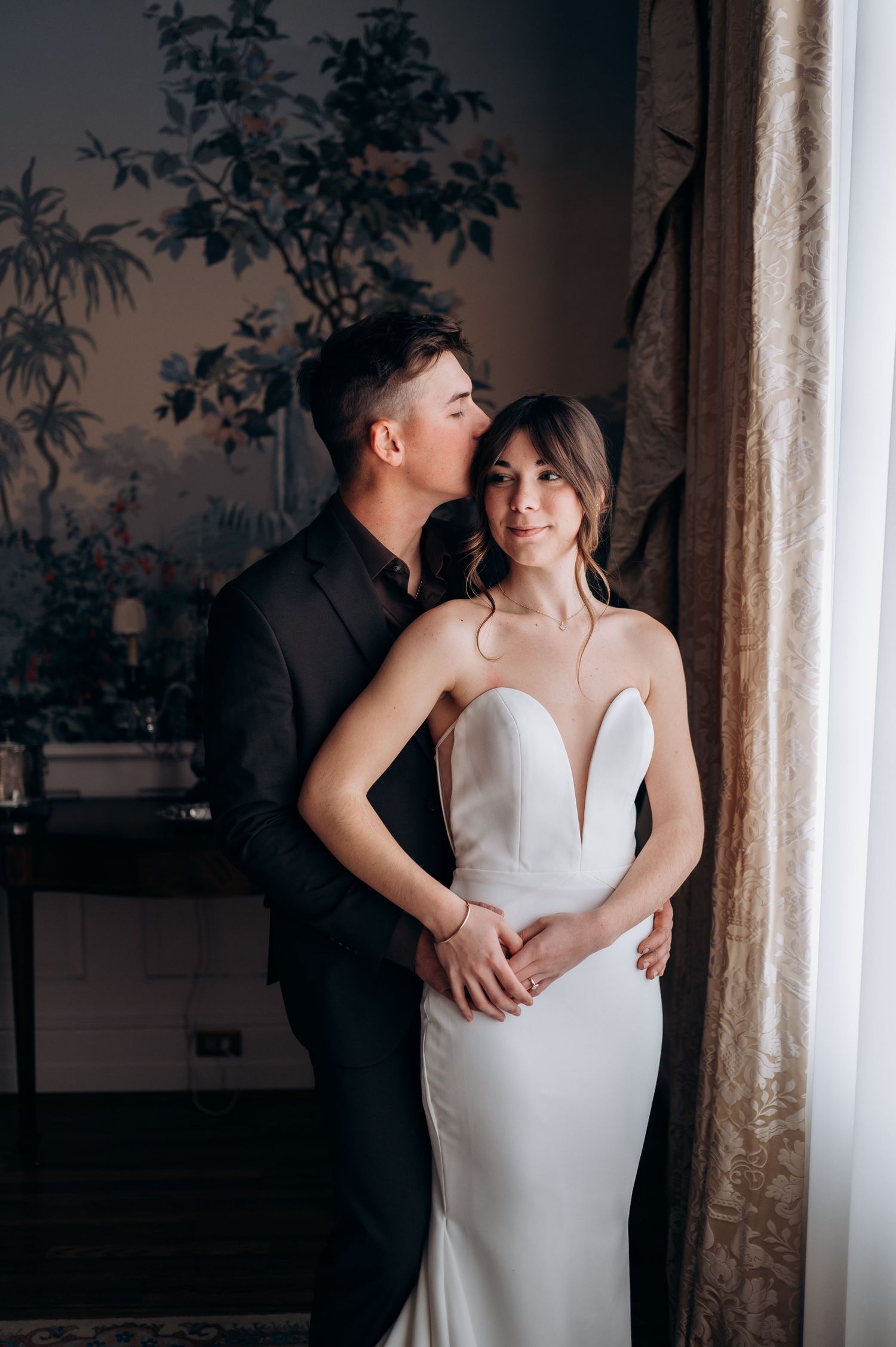 Groom in black suit kisses bride in sleek white gown by a window with floral wallpaper.
