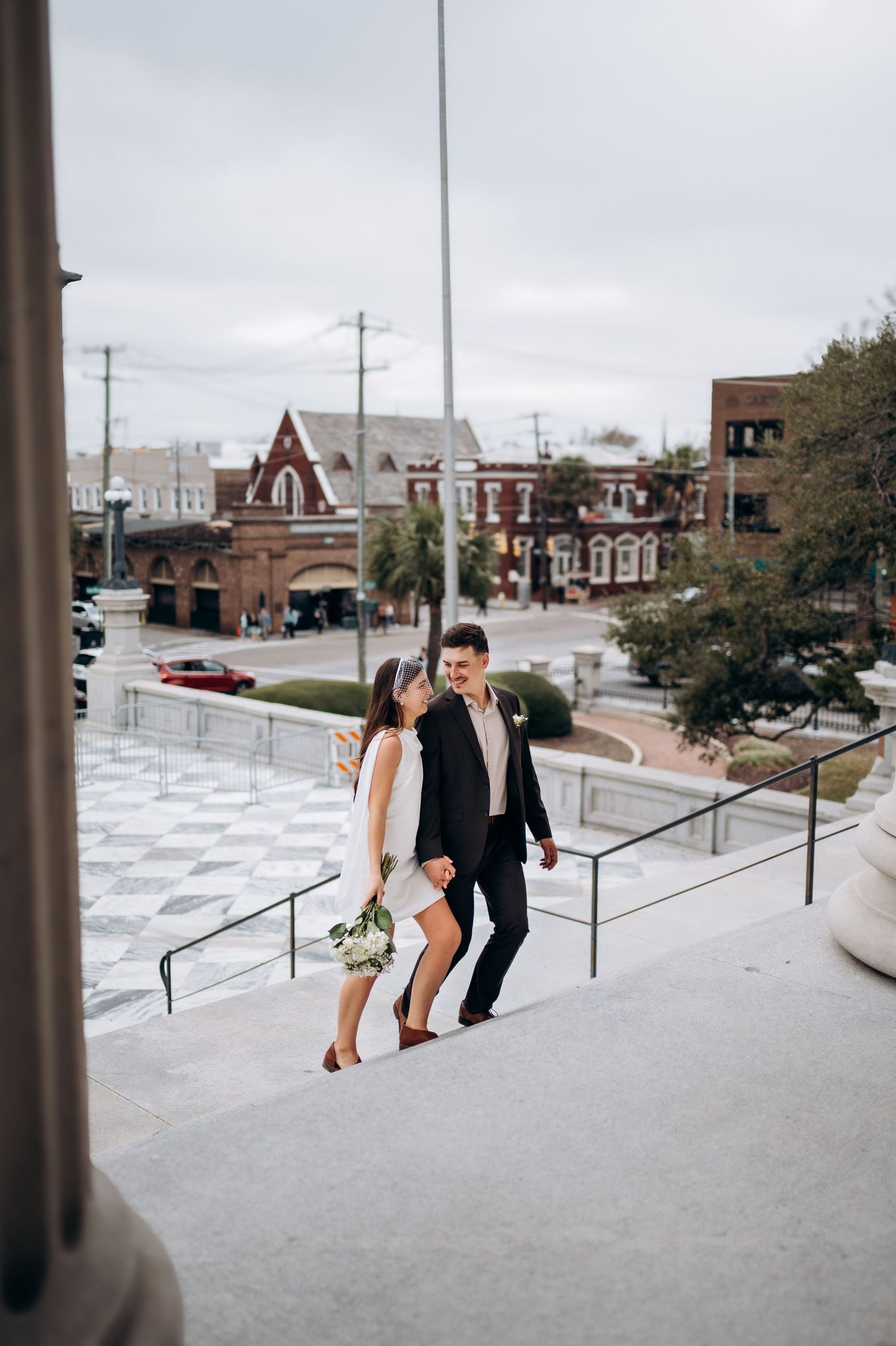 Wedding couple walks along outdoor terrace steps with urban cityscape backdrop on overcast day.