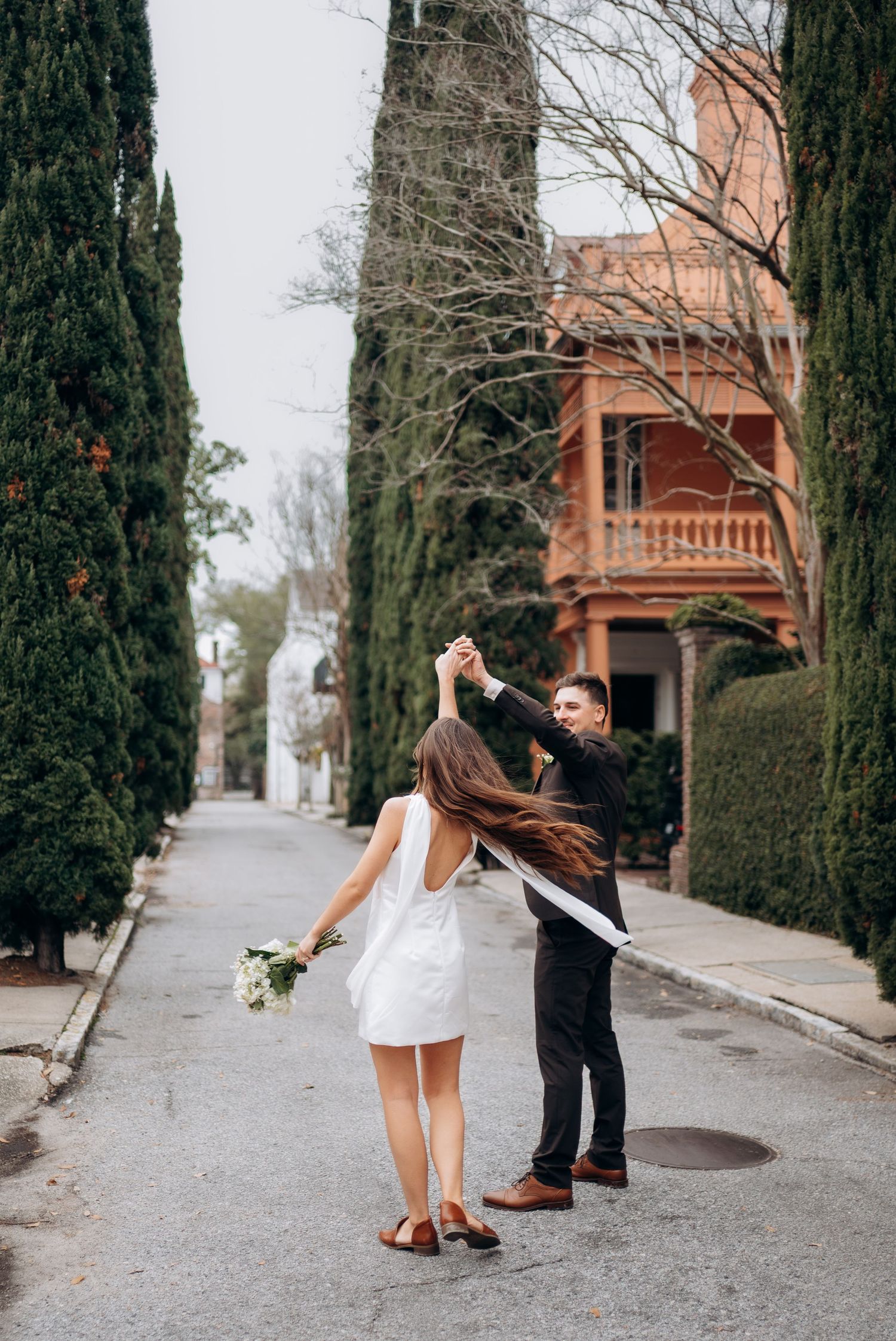 Groom spinning bride in white mini dress on scenic tree-lined street, her long hair flowing in the air.