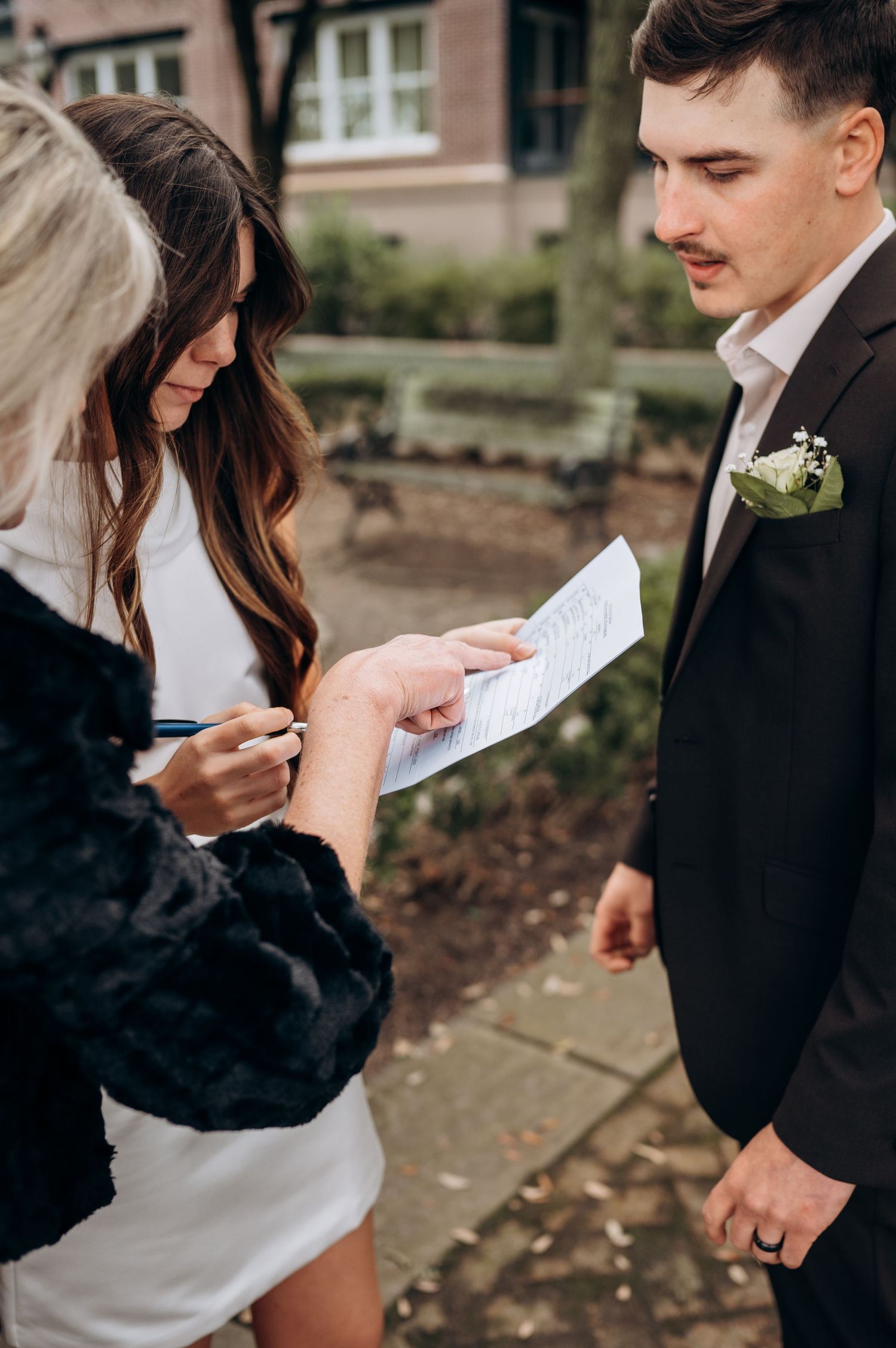 Officiant signing marriage certificate with bride in white dress and groom in dark suit with boutonniere.