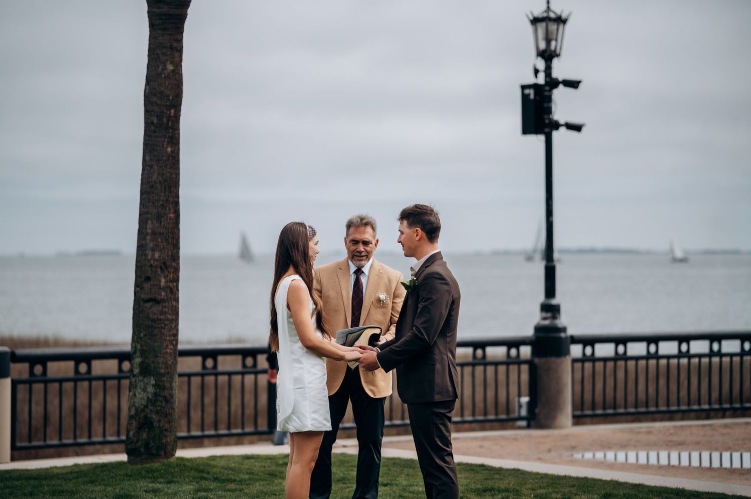 Couple exchanging vows with officiant by waterfront on cloudy day, outdoor elopement ceremony near lamp post.