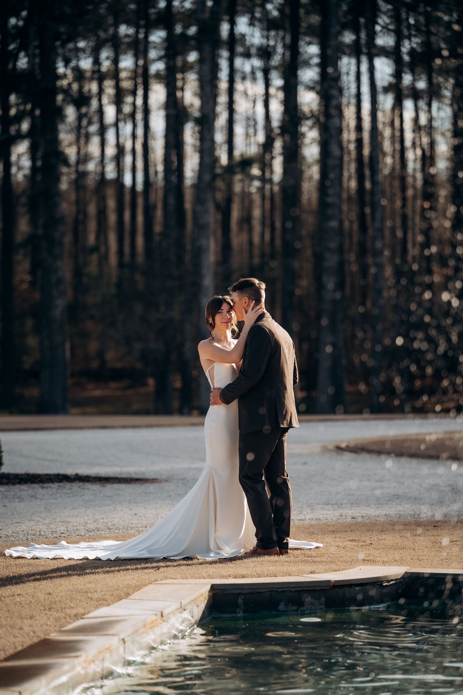 Bride and groom sharing romantic moment beside reflective pool with pine forest wedding backdrop.