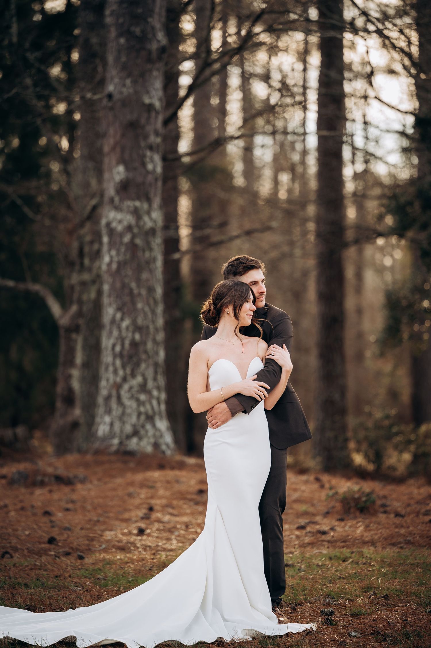 Bride in sleek white gown and groom in black suit share a tender embrace among tall pine trees.