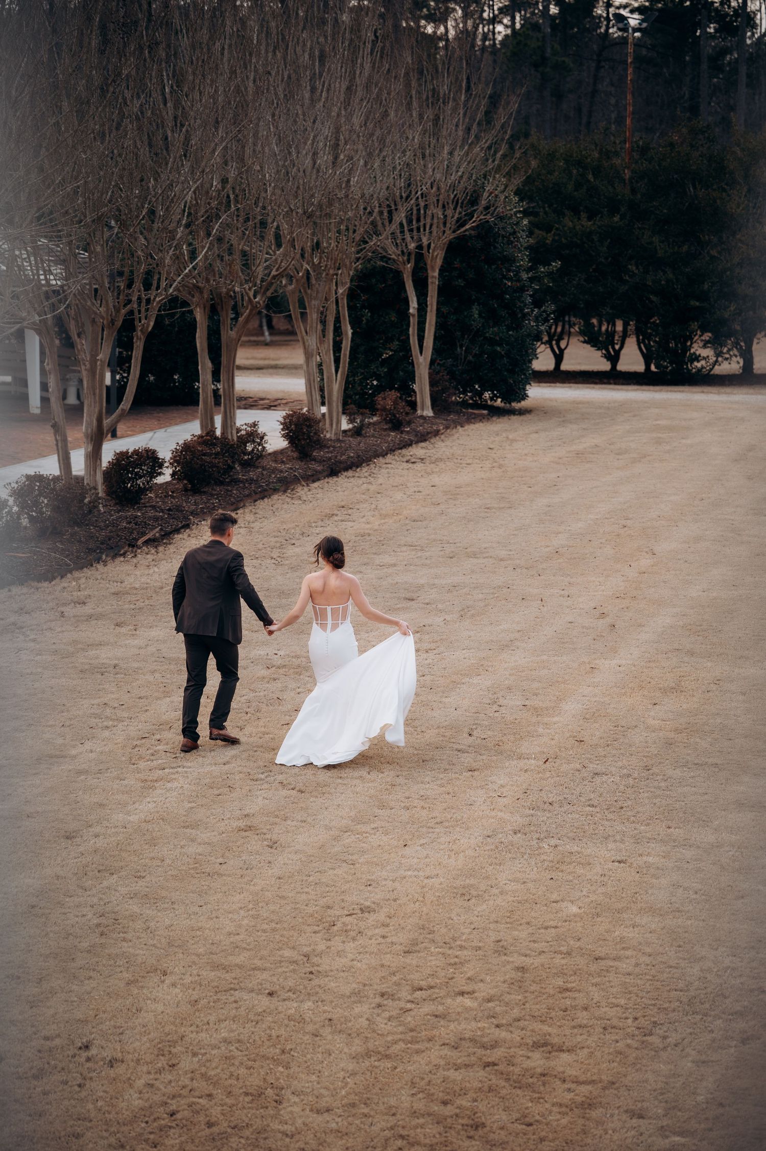 Bride and groom walk hand in hand across sandy grounds lined with bare winter trees at outdoor venue.