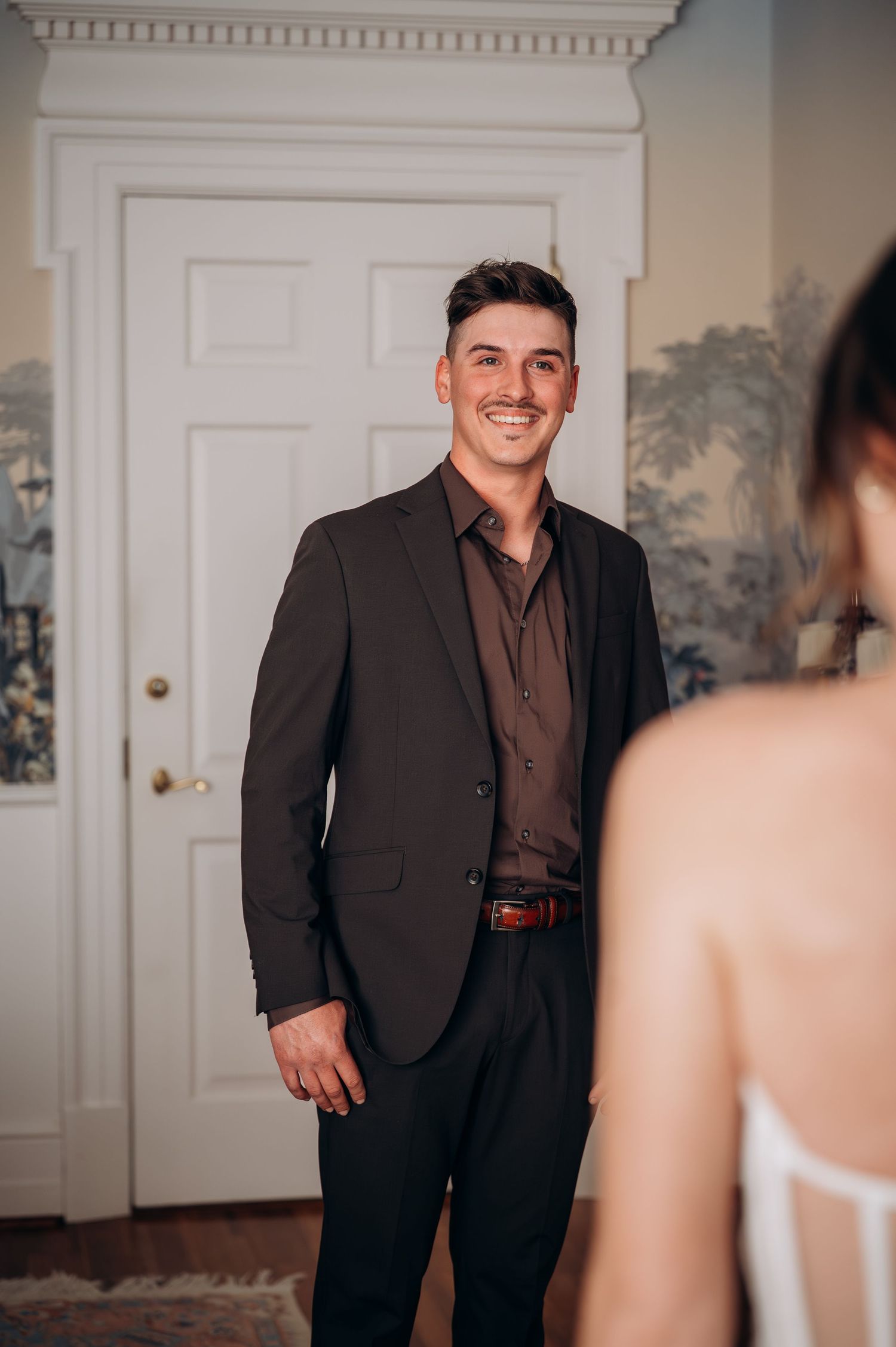 Smiling groom in dark suit sees bride for the first time in an elegant wallpapered room.