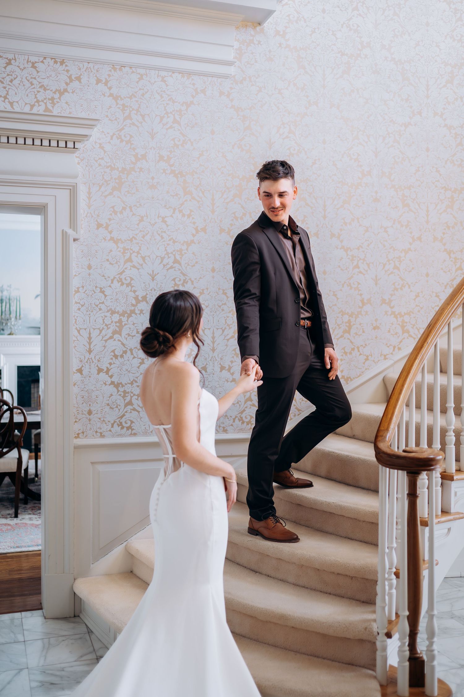 Smiling groom in black suit holds bride's hand on grand staircase with ornate white and gold wallpaper.
