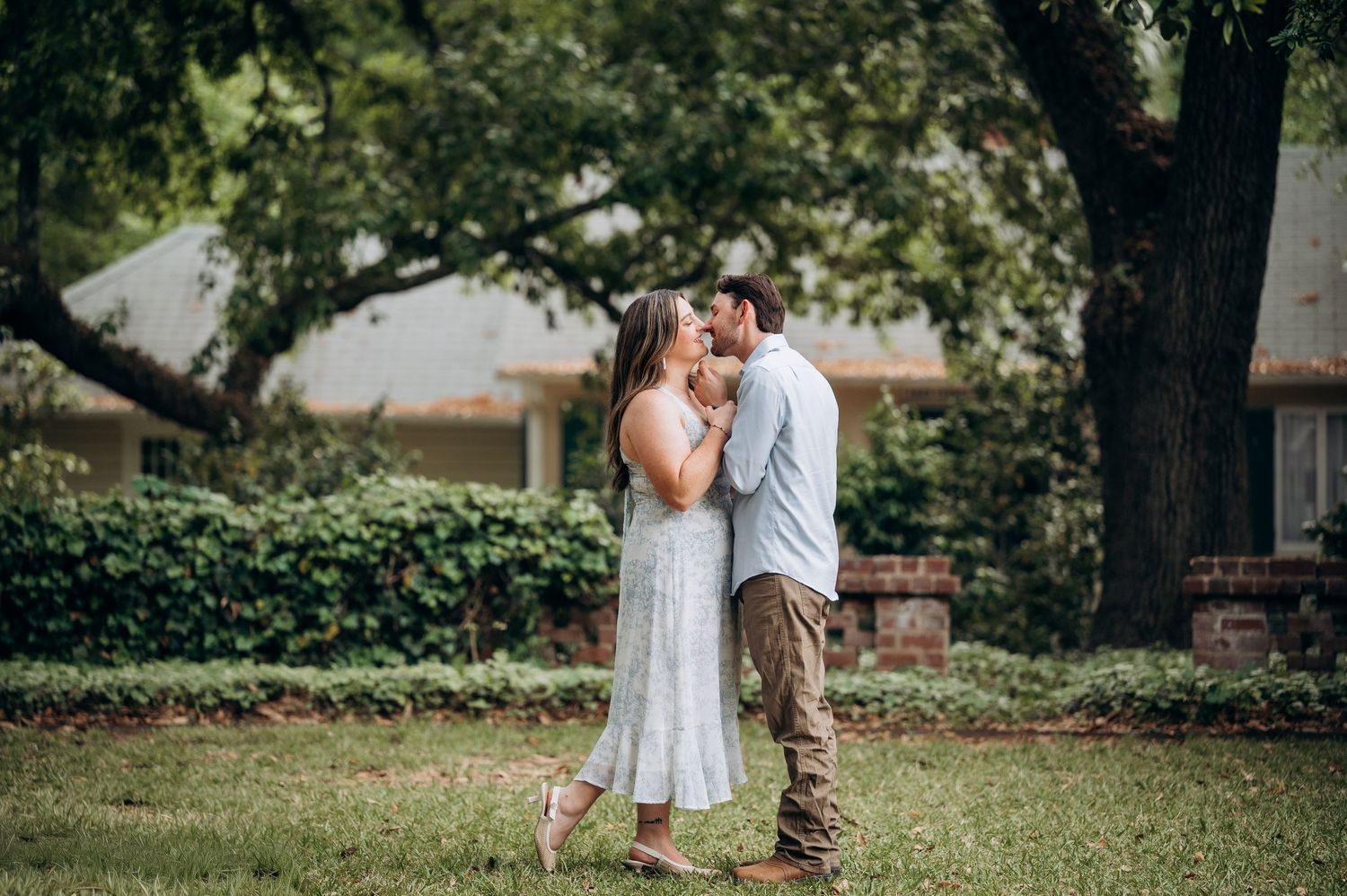 Couple sharing a tender kiss on a green lawn with trees and a brick building in the background.