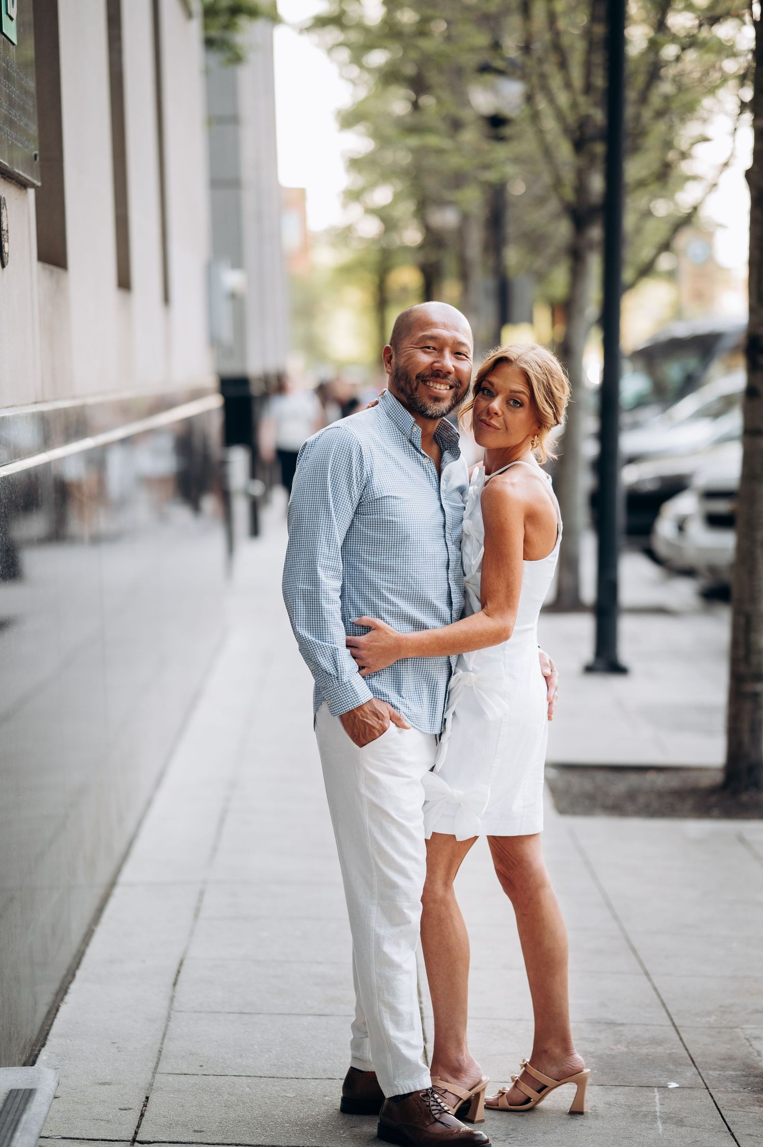 Happy couple hugging on urban sidewalk, man in blue shirt with white pants and woman in white mini dress.