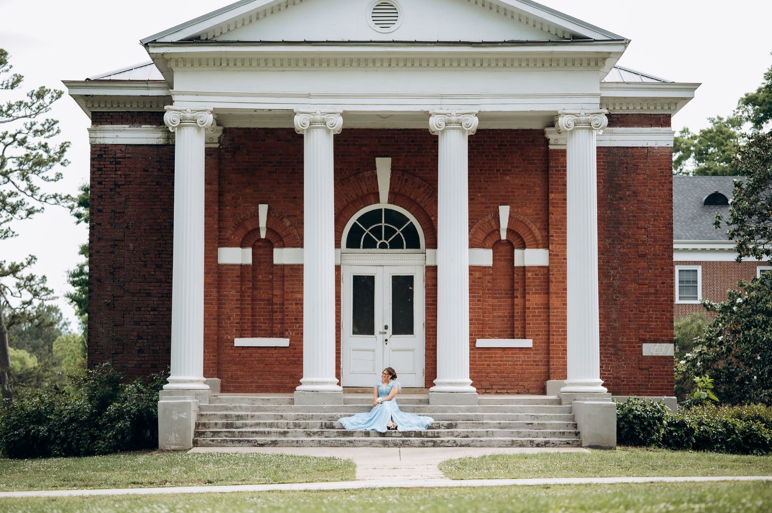 Woman in blue gown sitting on steps of classic brick building with white columns and arched window.
