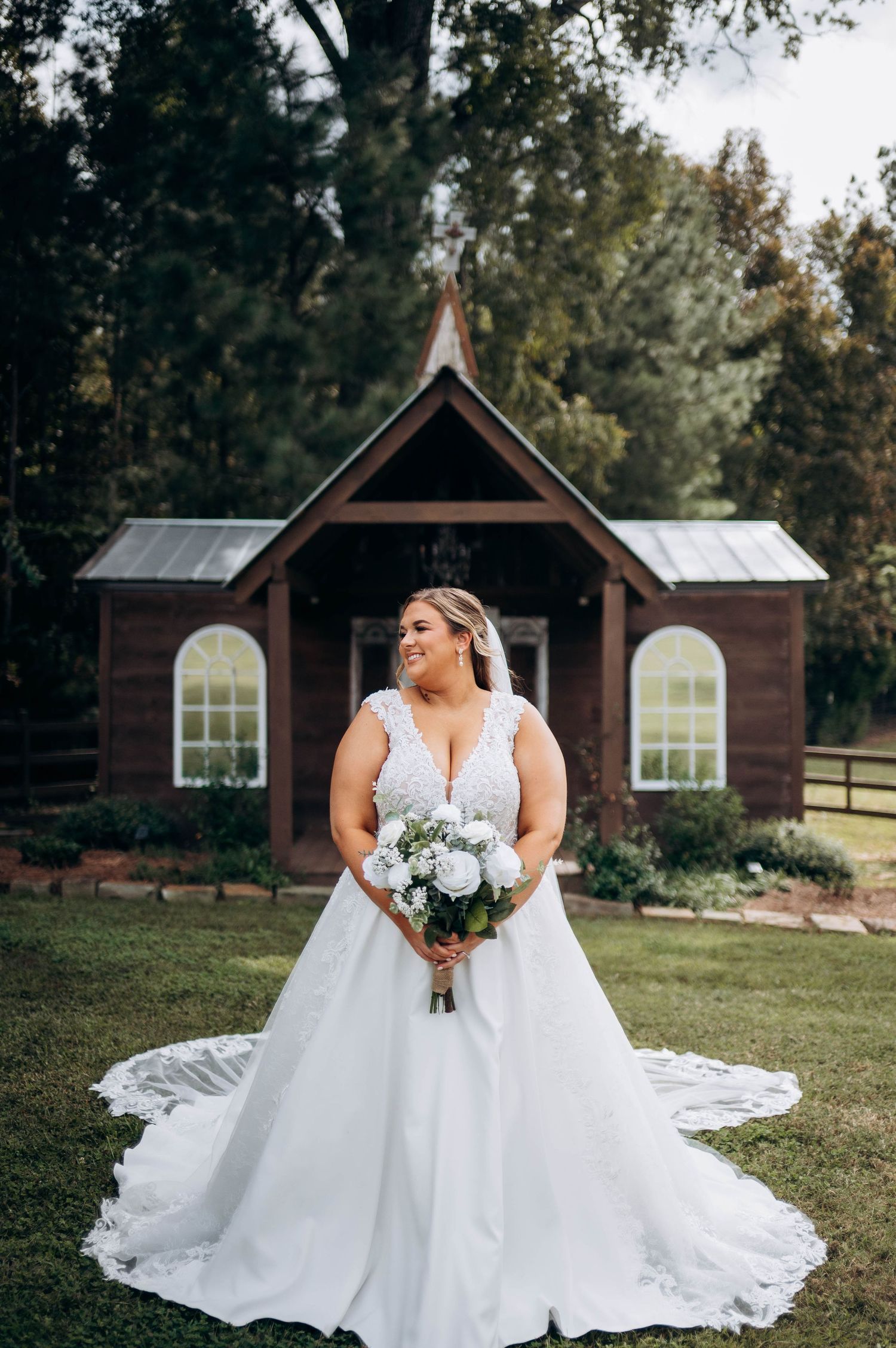 A bride in a flowing white lace gown poses in front of a rustic wooden chapel.