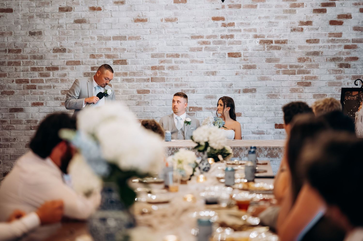 Wedding speaker toasts couple at elegant reception with brick wall backdrop and candlelit long tables.
