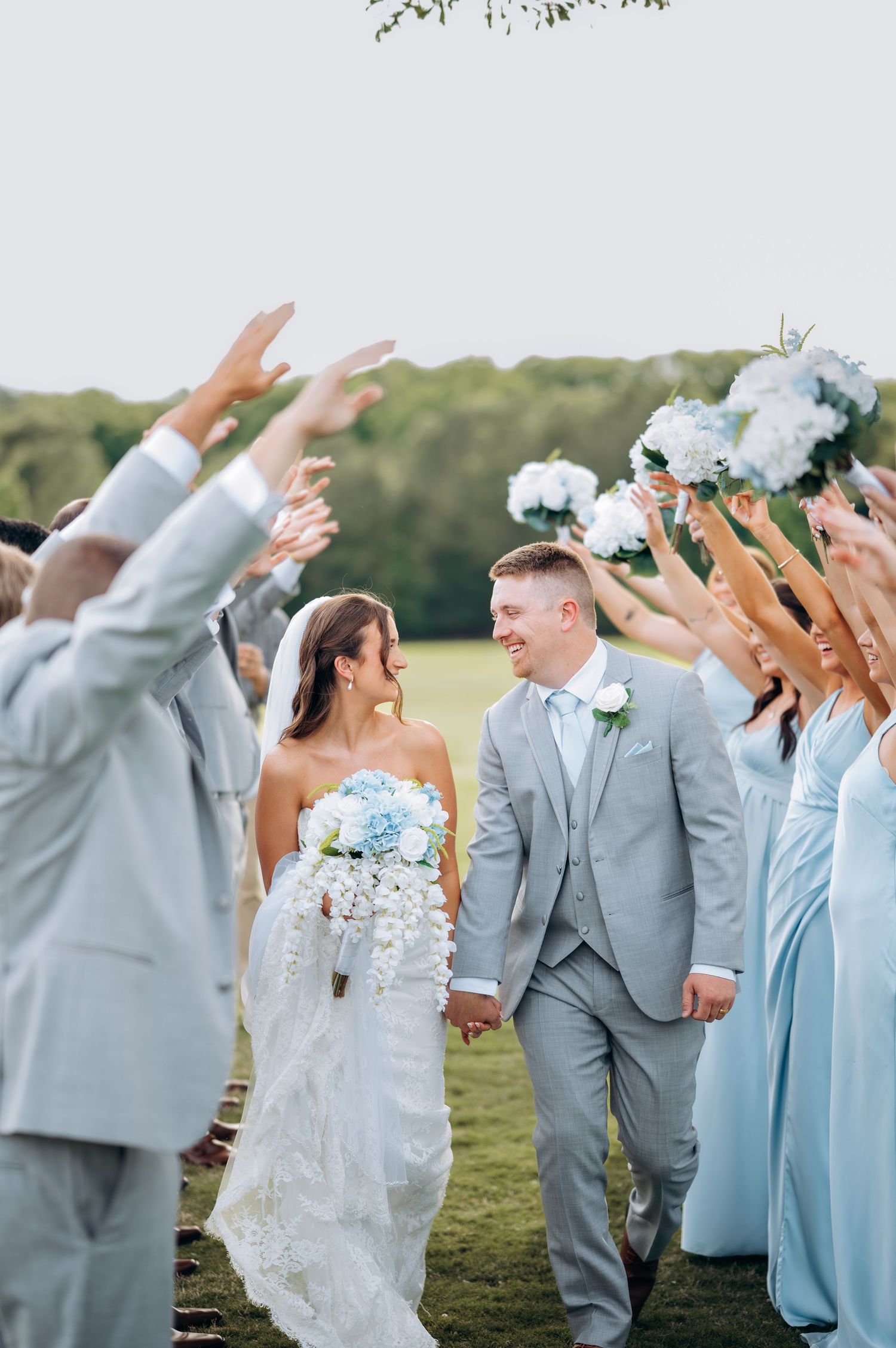 Bride and groom walk through cheering wedding party holding blue hydrangea bouquets outdoors.