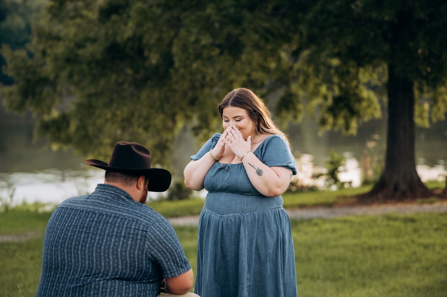 Emotional woman covering her mouth during a marriage proposal in a park, man kneeling before her in a black cowboy hat.