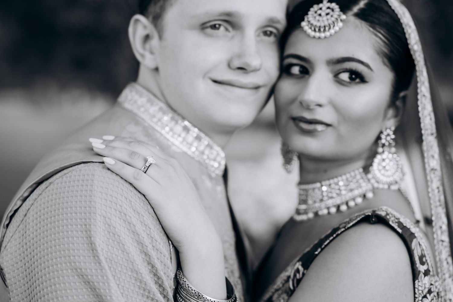 Black and white portrait of an interracial couple in traditional Indian wedding attire, smiling closely together.
