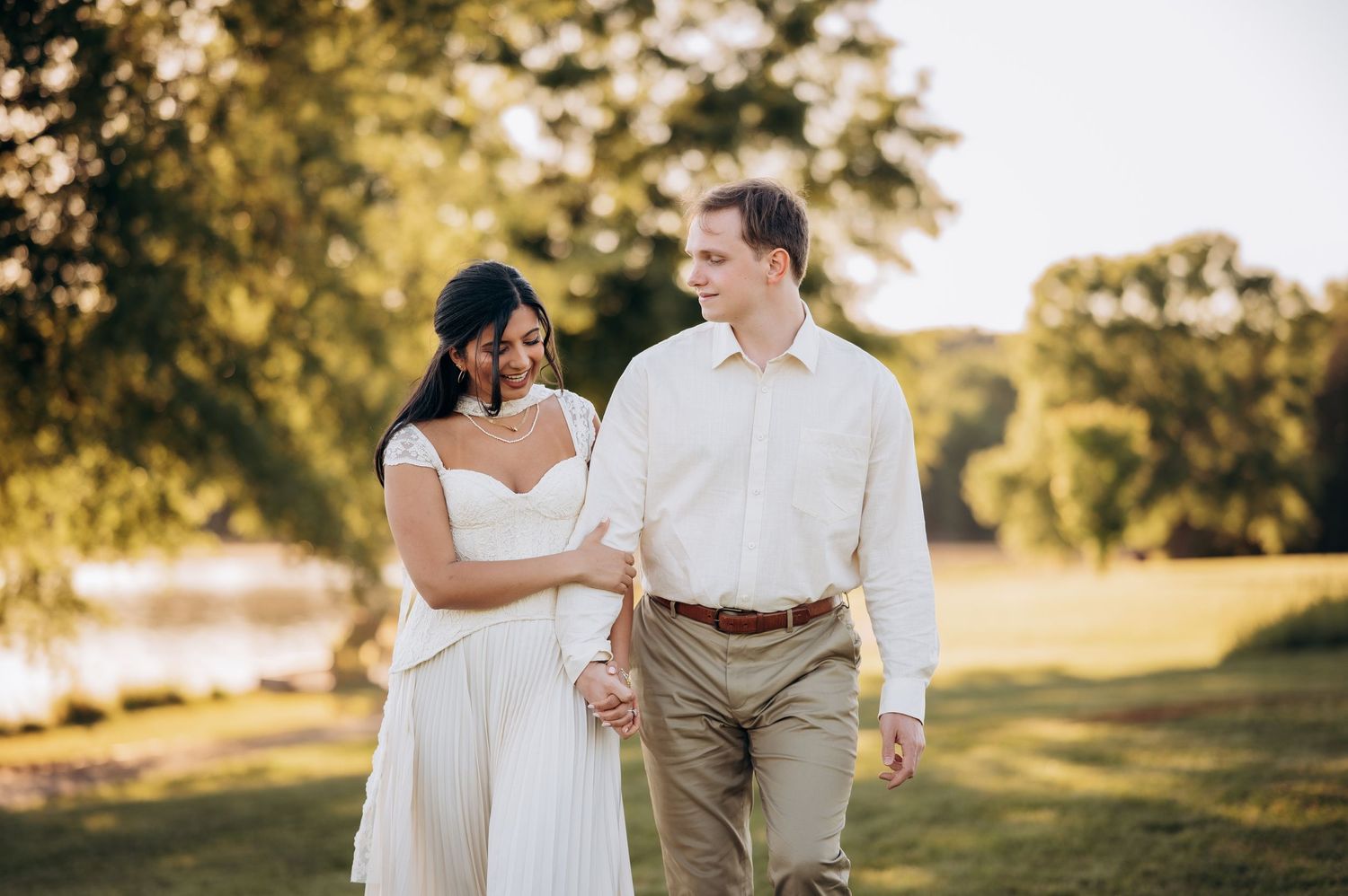 Couple walks hand-in-hand outdoors in golden hour light, woman in white dress, man in khakis, trees and lake behind them.