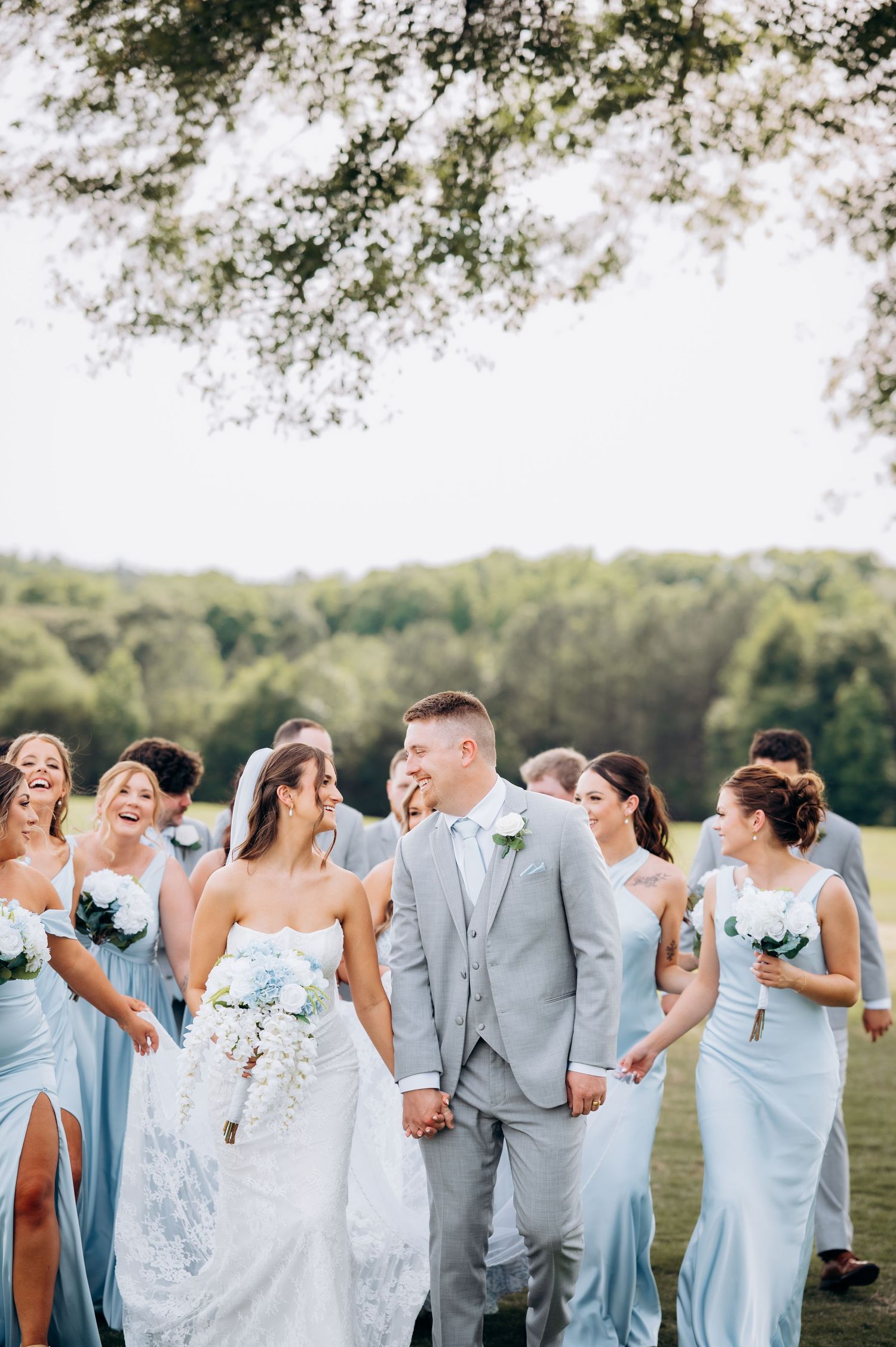 A joyful bride and groom hold hands surrounded by bridesmaids in light blue dresses and groomsmen in gray suits outdoors.