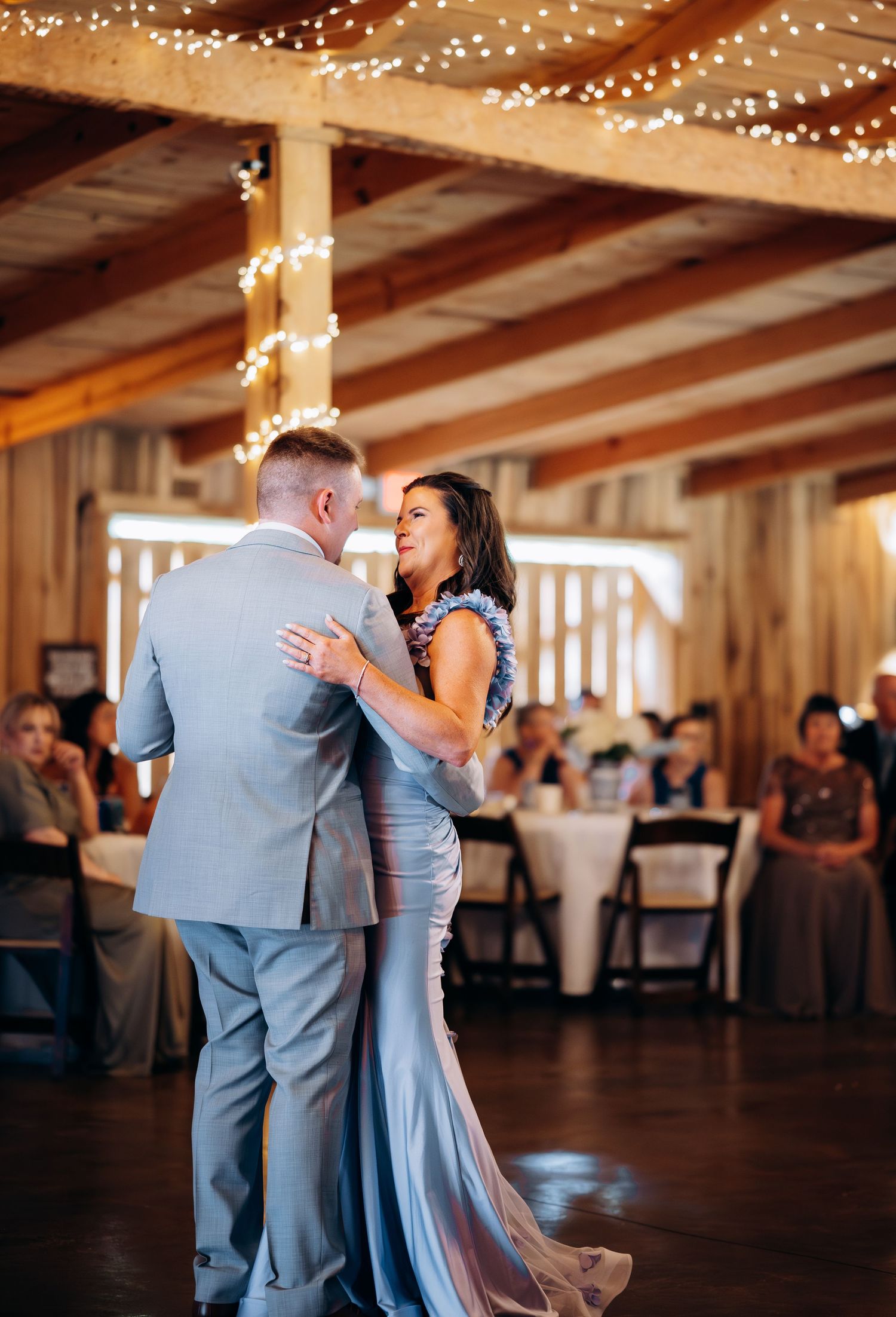 Couple shares first dance in rustic barn wedding venue with twinkling fairy lights and guests seated in background.
