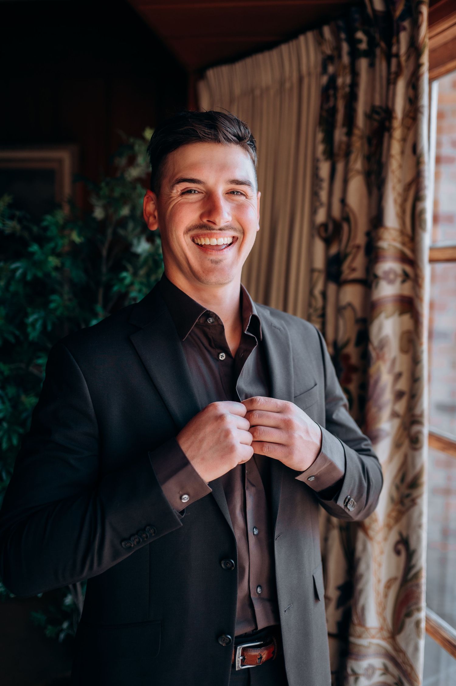 Groom in black suit adjusts his collar while smiling in an elegant indoor setting.
