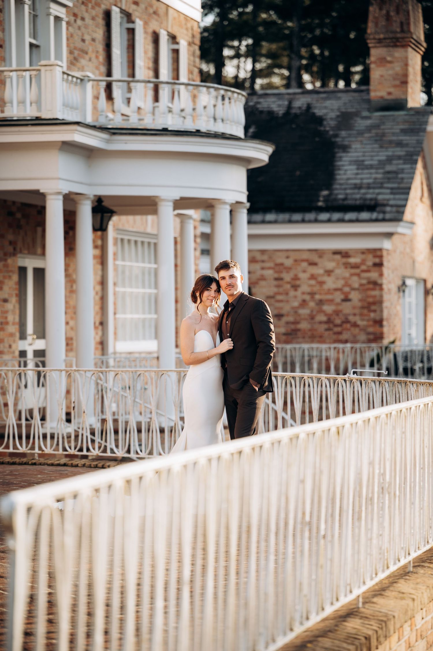 Couple in wedding attire poses along a white railing at a grand colonial-style venue.