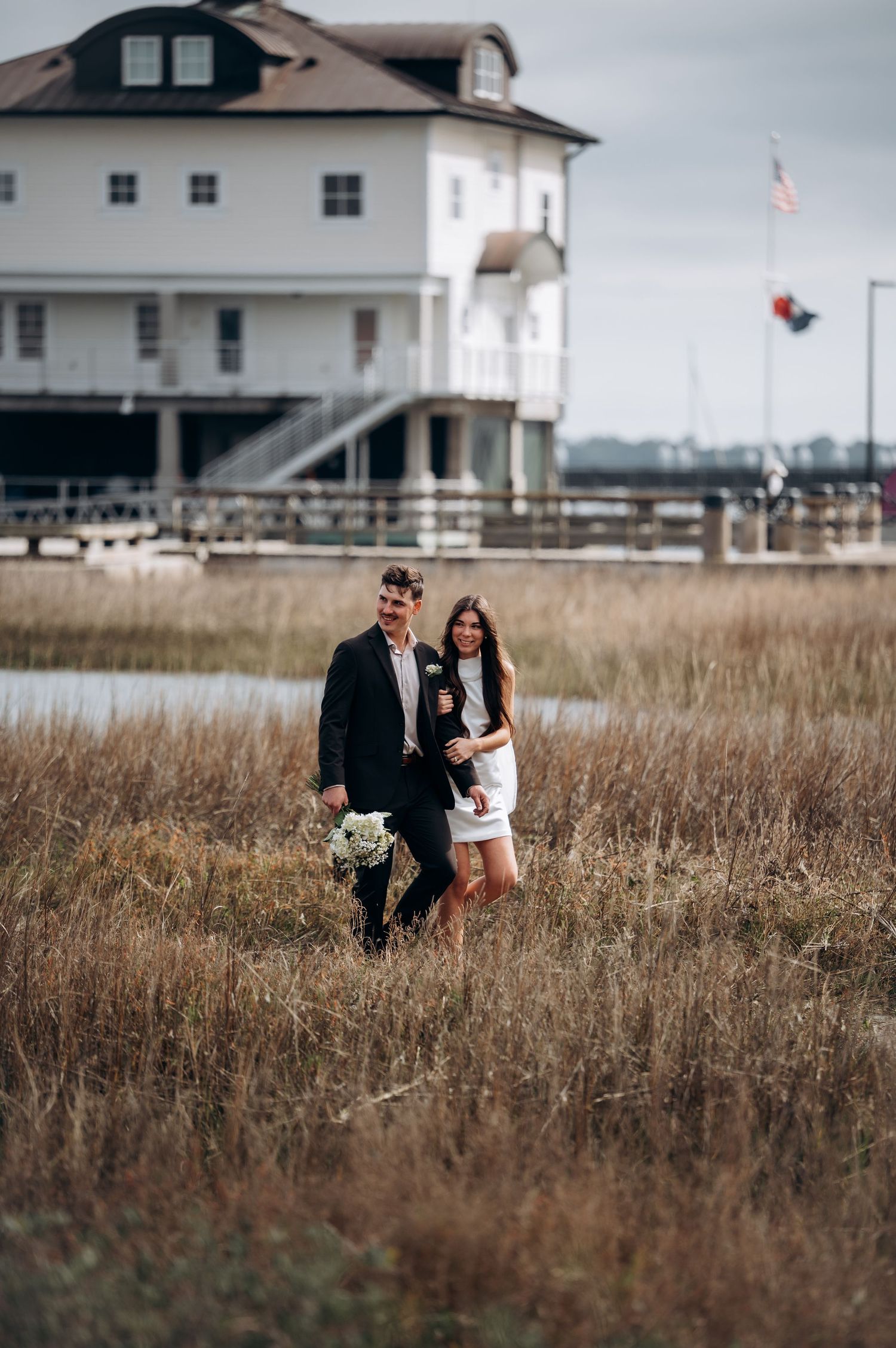 Couple walks through tall grass with coastal houses in background.