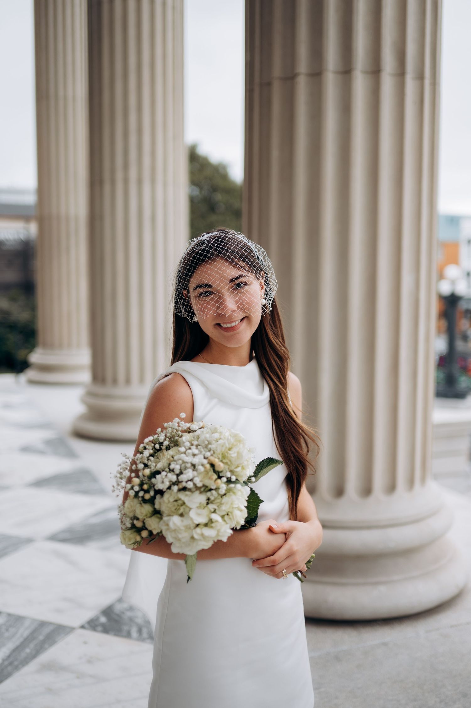 A bride in a classic white dress holds a bouquet of white hydrangeas while posing between elegant columns.