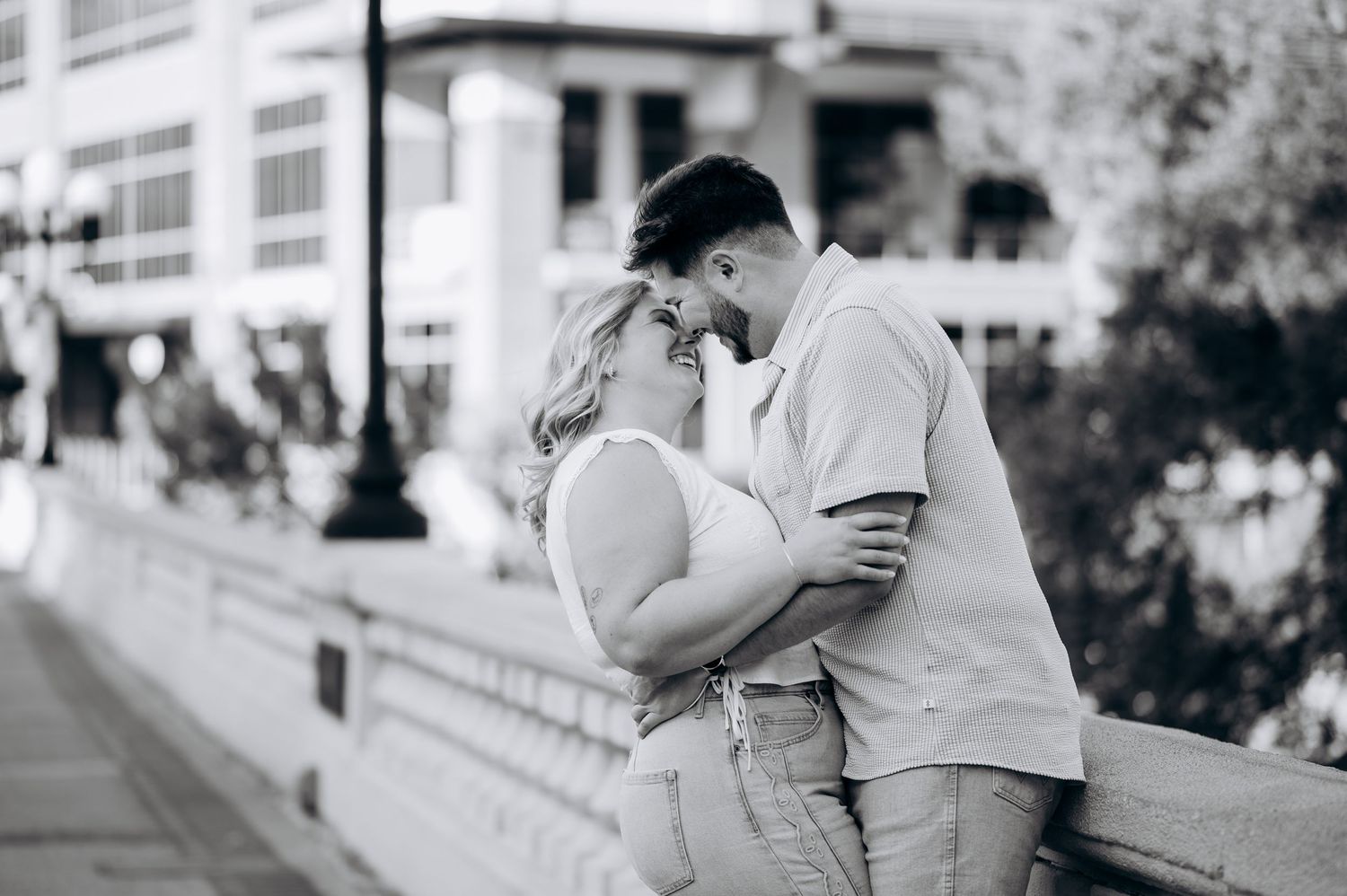 Black and white photo of a romantic couple leaning against a bridge railing, gazing into each other's eyes.