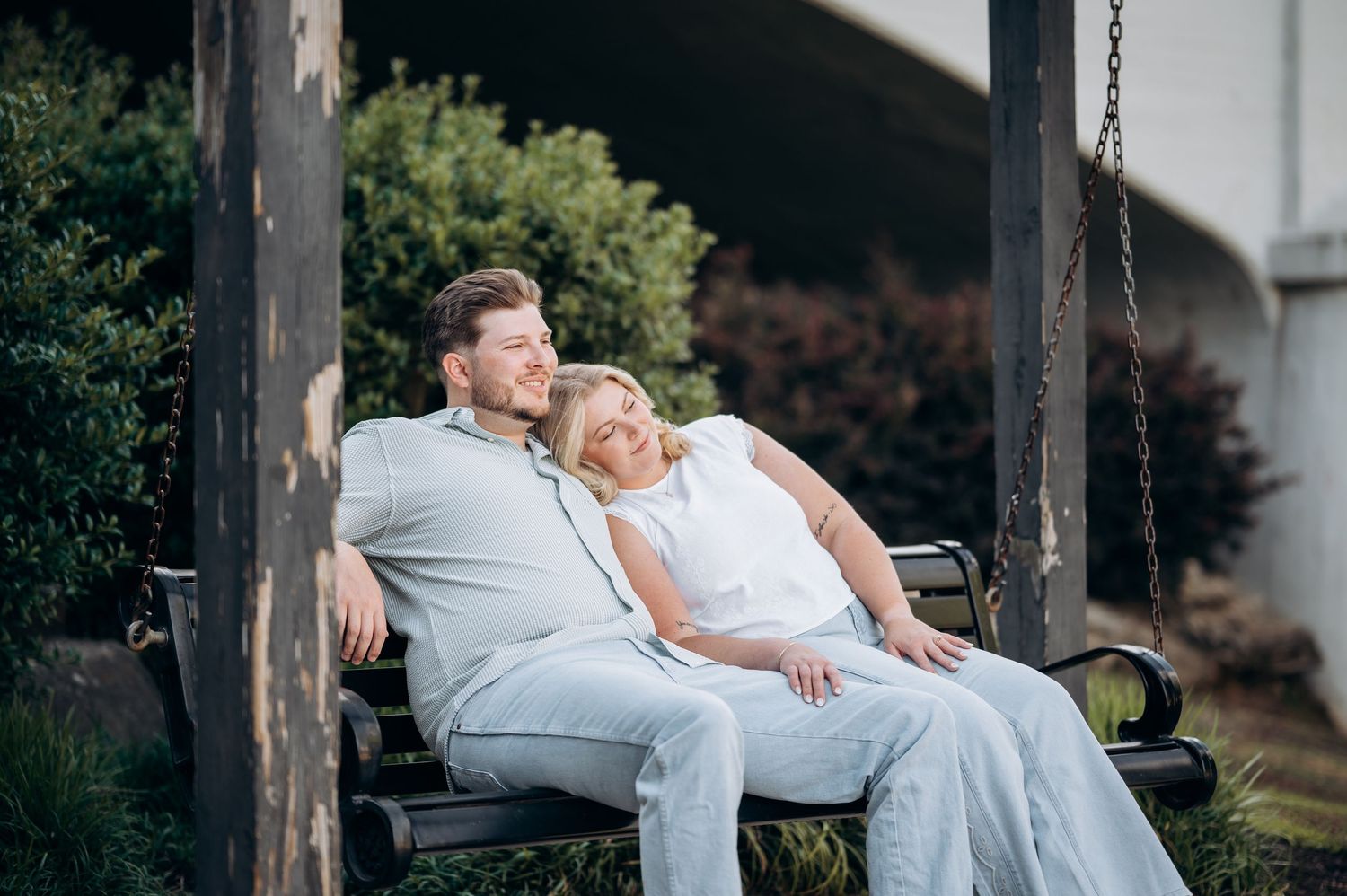 Couple relaxing on a wooden chain swing bench outdoors, woman leaning on man's shoulder in a garden setting.