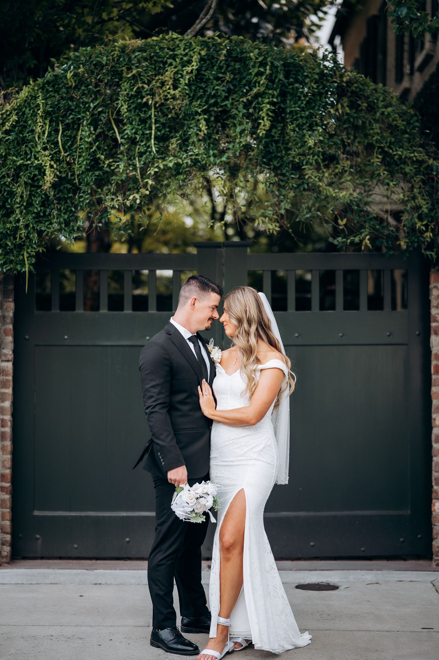 Bride and groom share romantic moment before dark gate covered in lush green ivy.