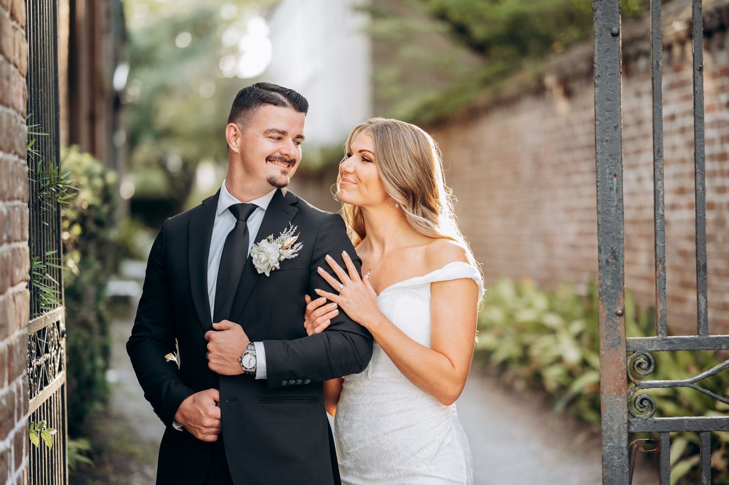 Bride and groom pose elegantly outdoors, she in white off-shoulder gown, he in black suit with floral boutonniere.