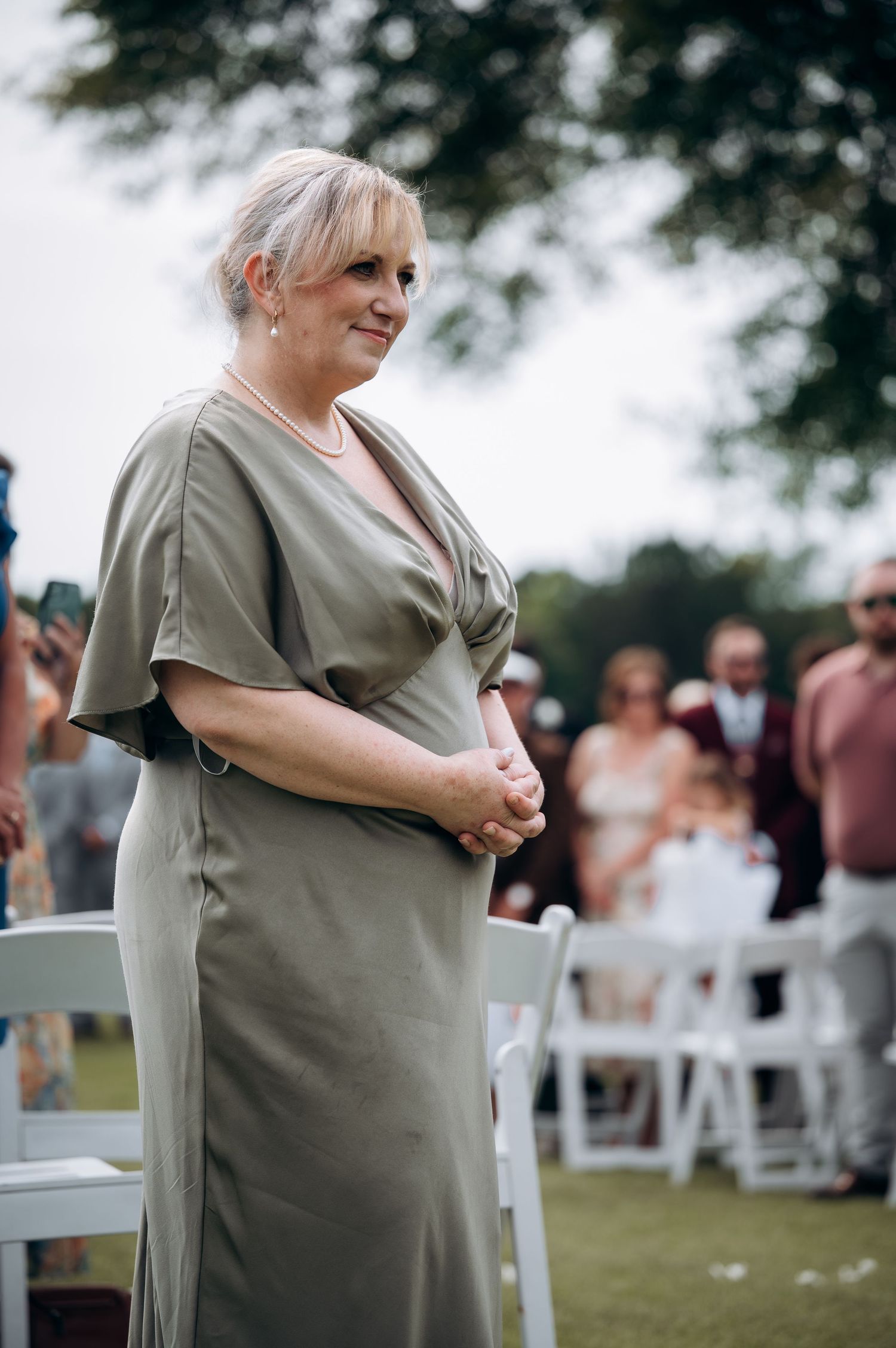 Emotional mother of bride in olive green dress watching outdoor wedding ceremony.
