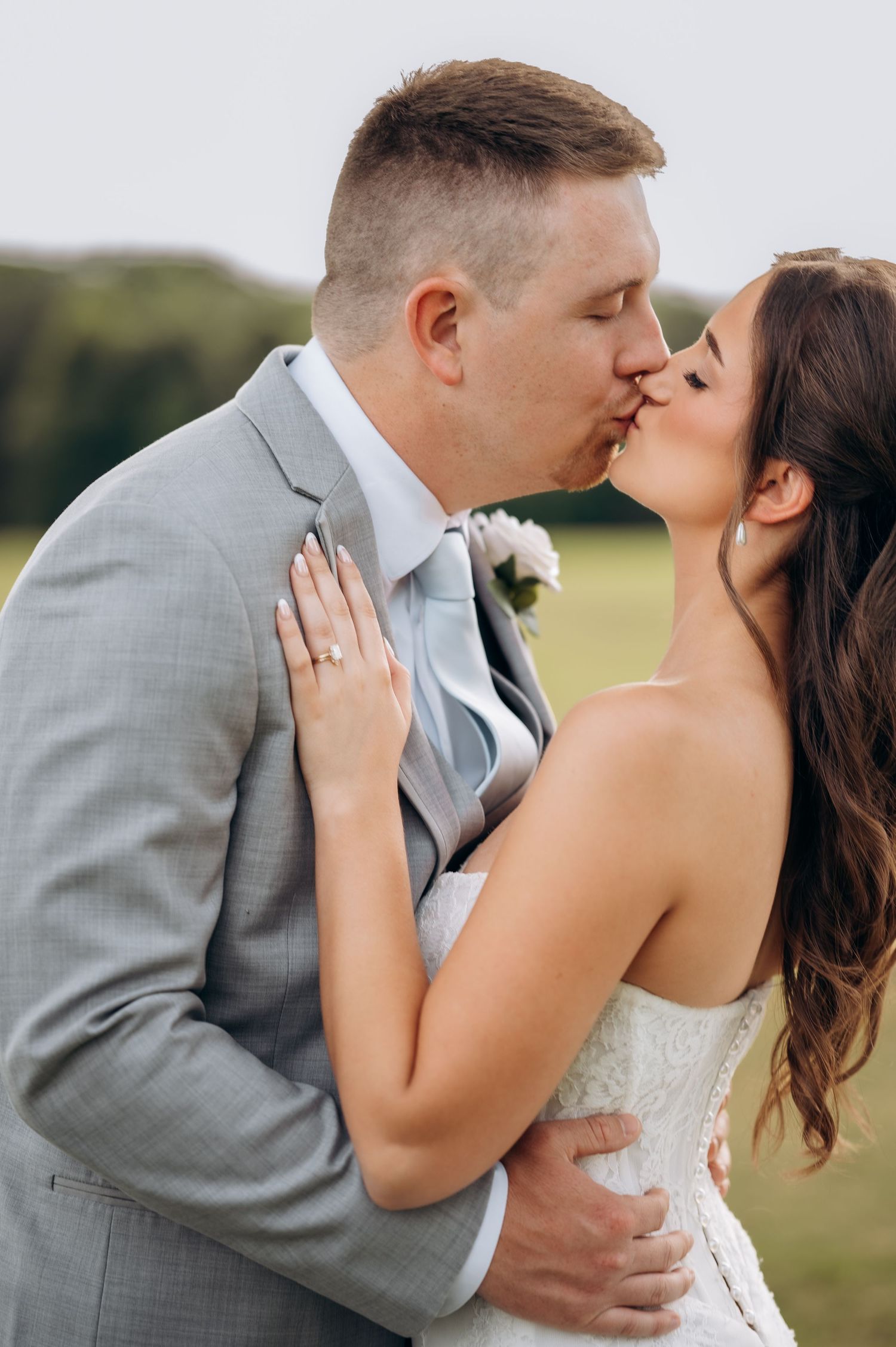 Bride and groom share a romantic kiss outdoors on their wedding day, with lush green fields in the background.