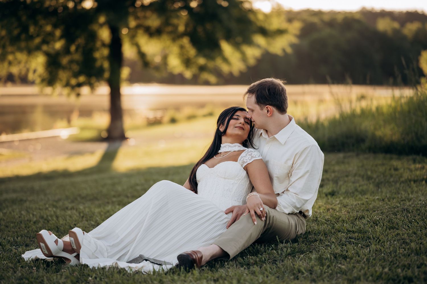 Couple sitting on lush green grass near a lake at golden hour, woman in white dress leaning against man in white shirt.