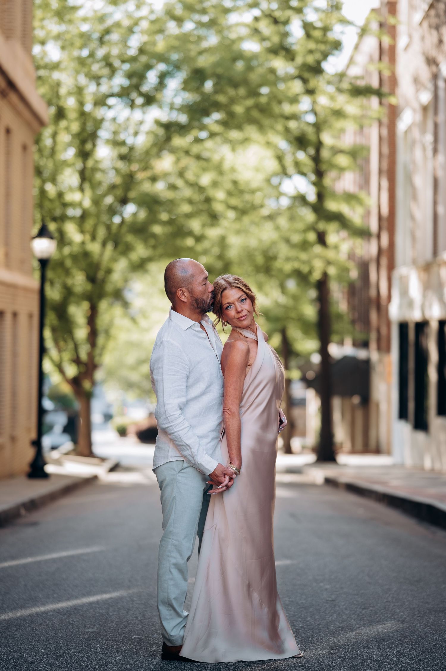 A couple shares a romantic embrace on a quiet urban street, the woman in a elegant blush gown and man in light linen attire.