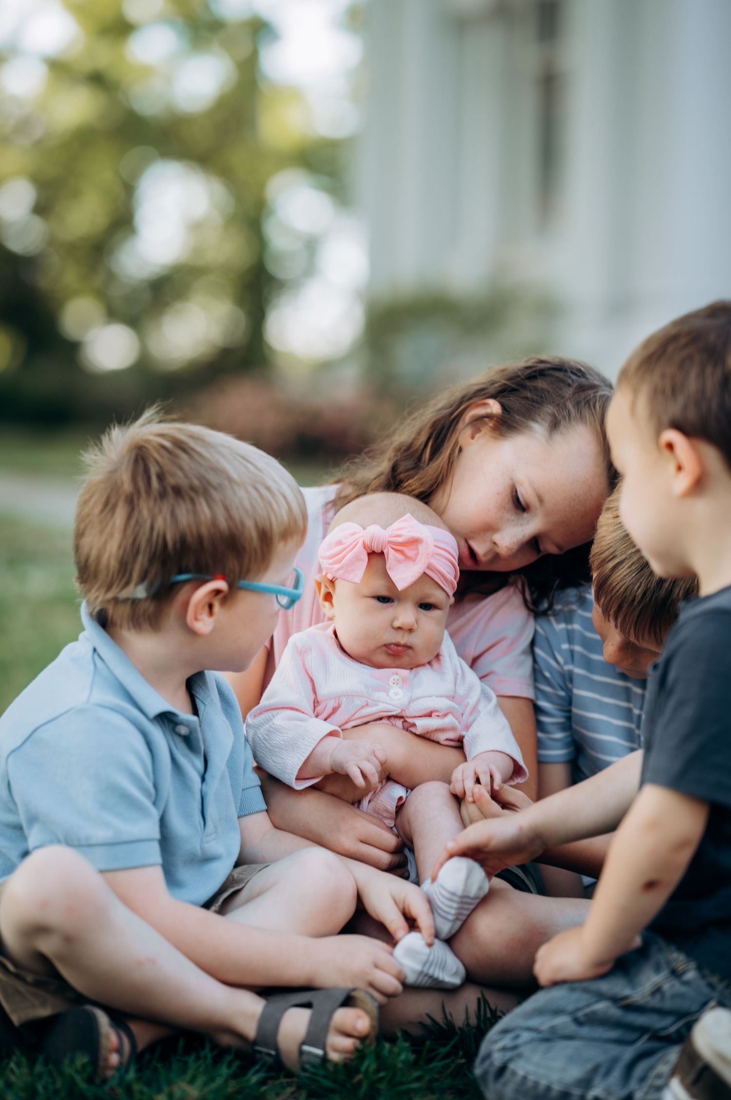 Four young siblings sit together on grass, older girl holding a baby in a pink bow while brothers look on adoringly.