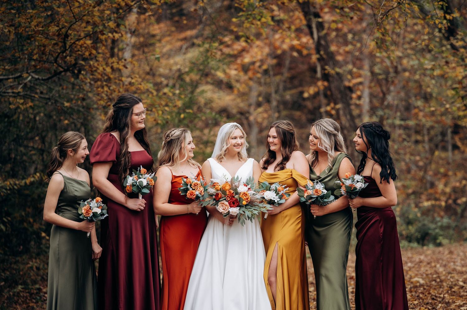 Bridal party poses together in autumn forest wearing burgundy, yellow and white dresses.