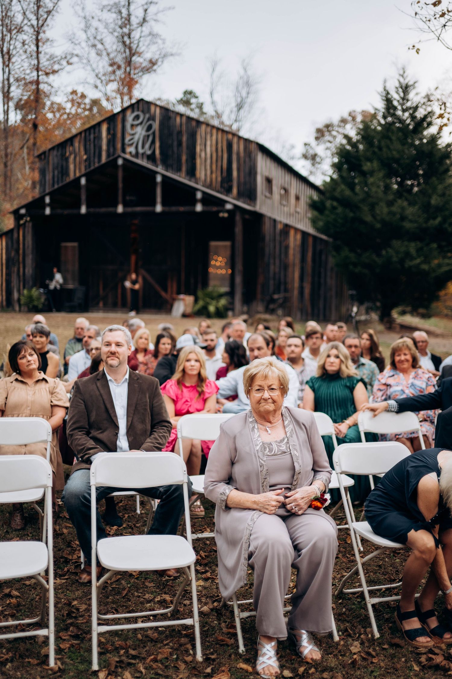 Wedding guests seated in white folding chairs at outdoor ceremony with rustic barn backdrop.