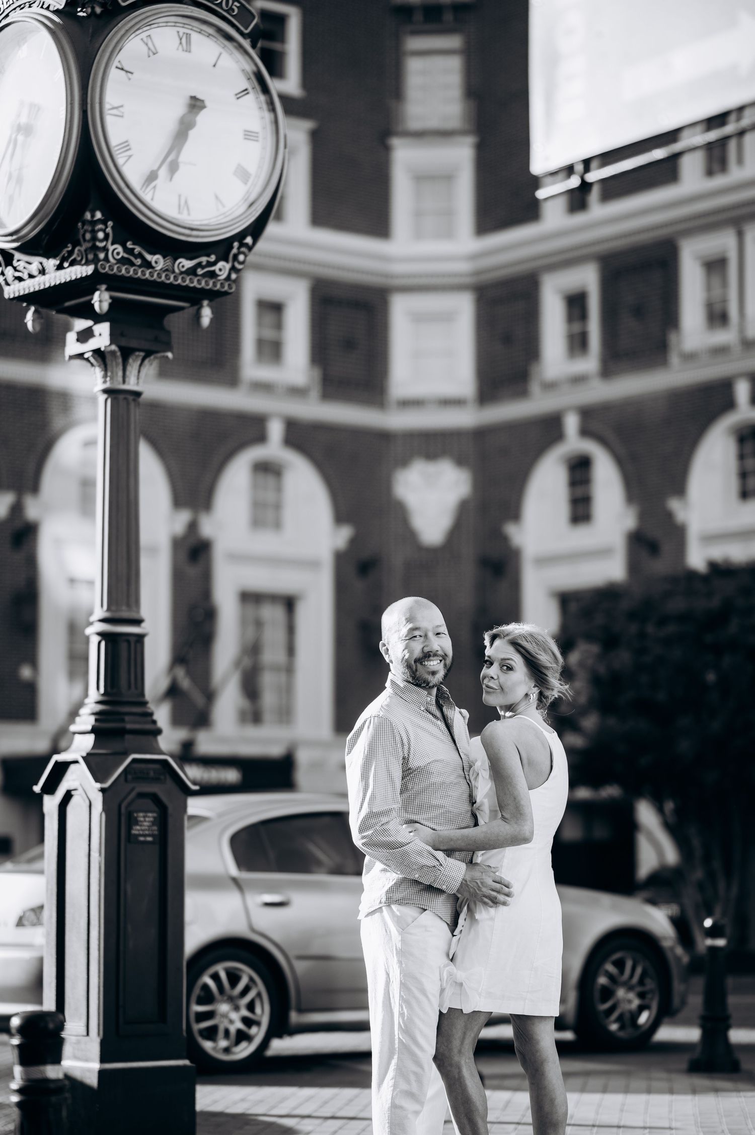 Black and white photo of a couple embracing near a decorative street clock in an urban setting.