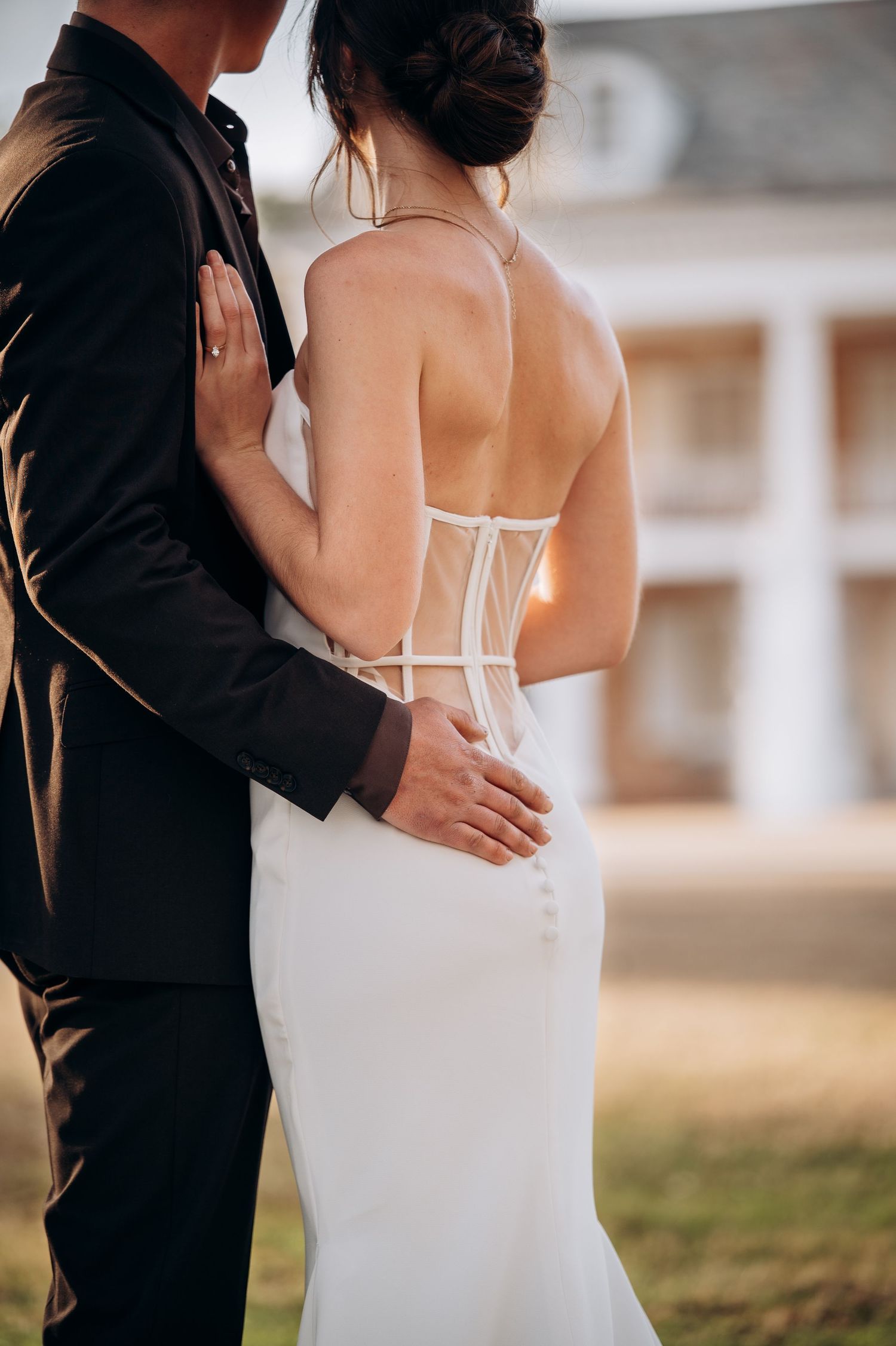 Close-up of bride's open corset back wedding gown as groom places hand on her waist outdoors.