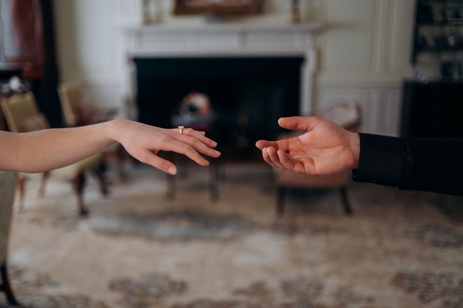 Bride and groom reaching toward each other showing engagement ring near elegant fireplace.