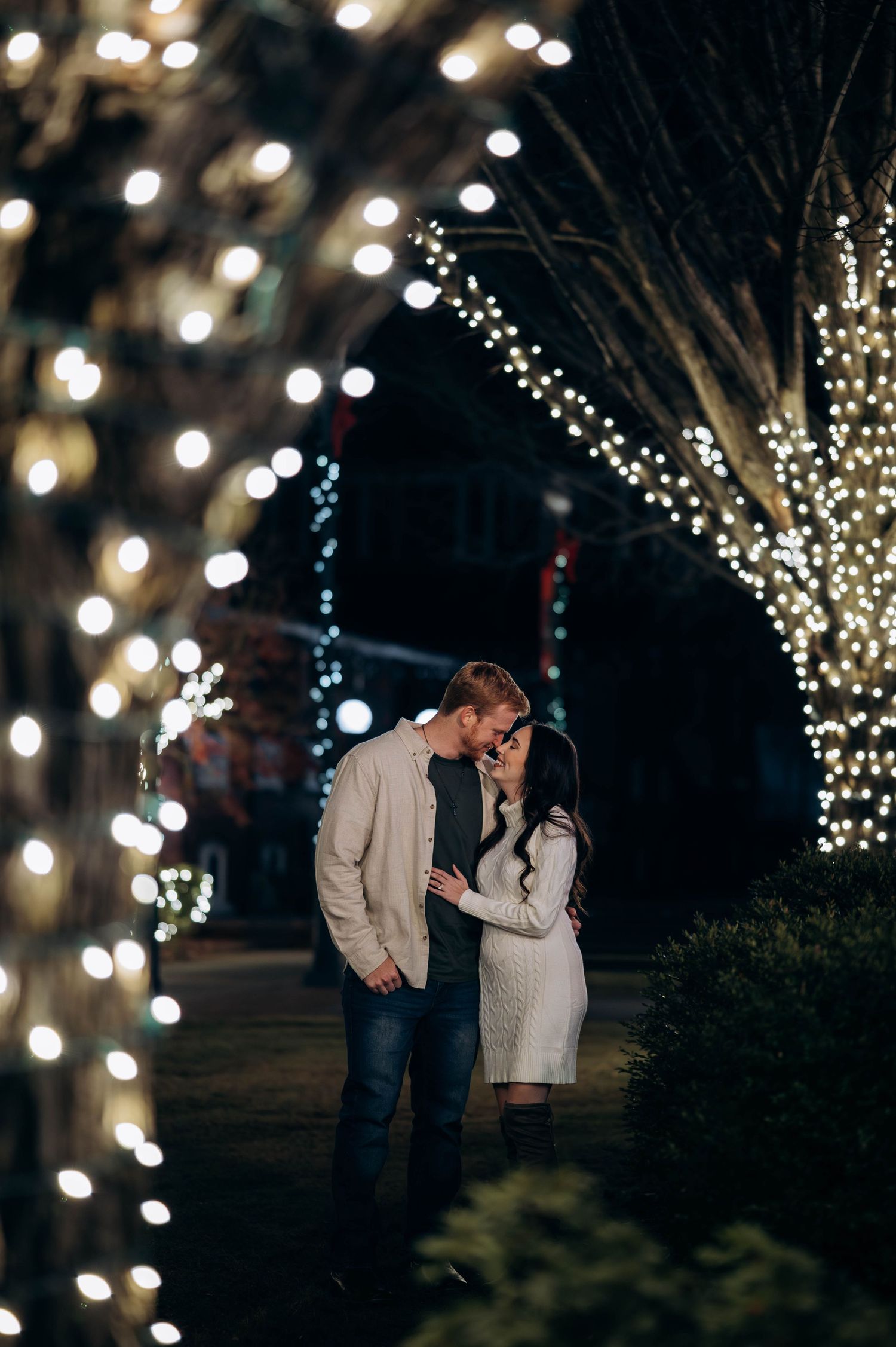 Couple sharing a romantic moment under twinkling fairy lights wrapped around trees at night.