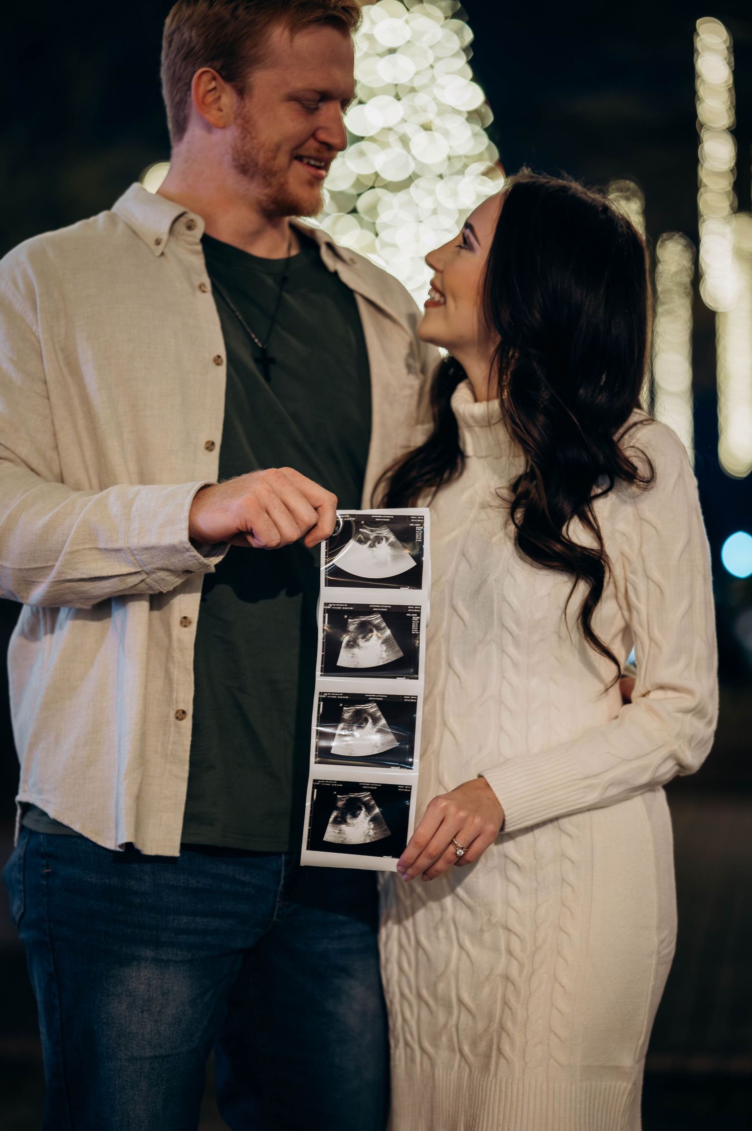 Couple smiling at each other holding ultrasound photos in front of a Christmas tree for a pregnancy announcement.