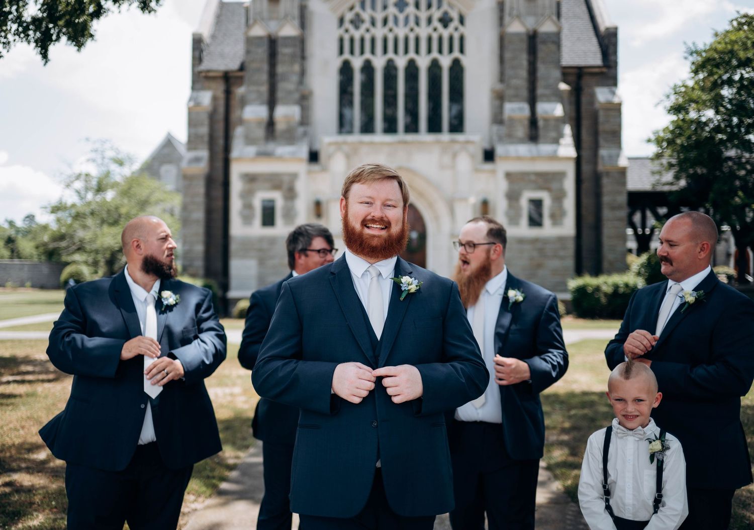 Wedding party gathered outside a gothic stone church with groomsmen in navy suits.