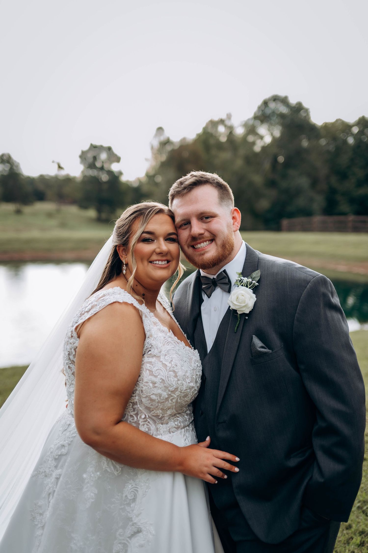 A romantic wedding portrait of a couple in formal attire standing by a lake at sunset.
