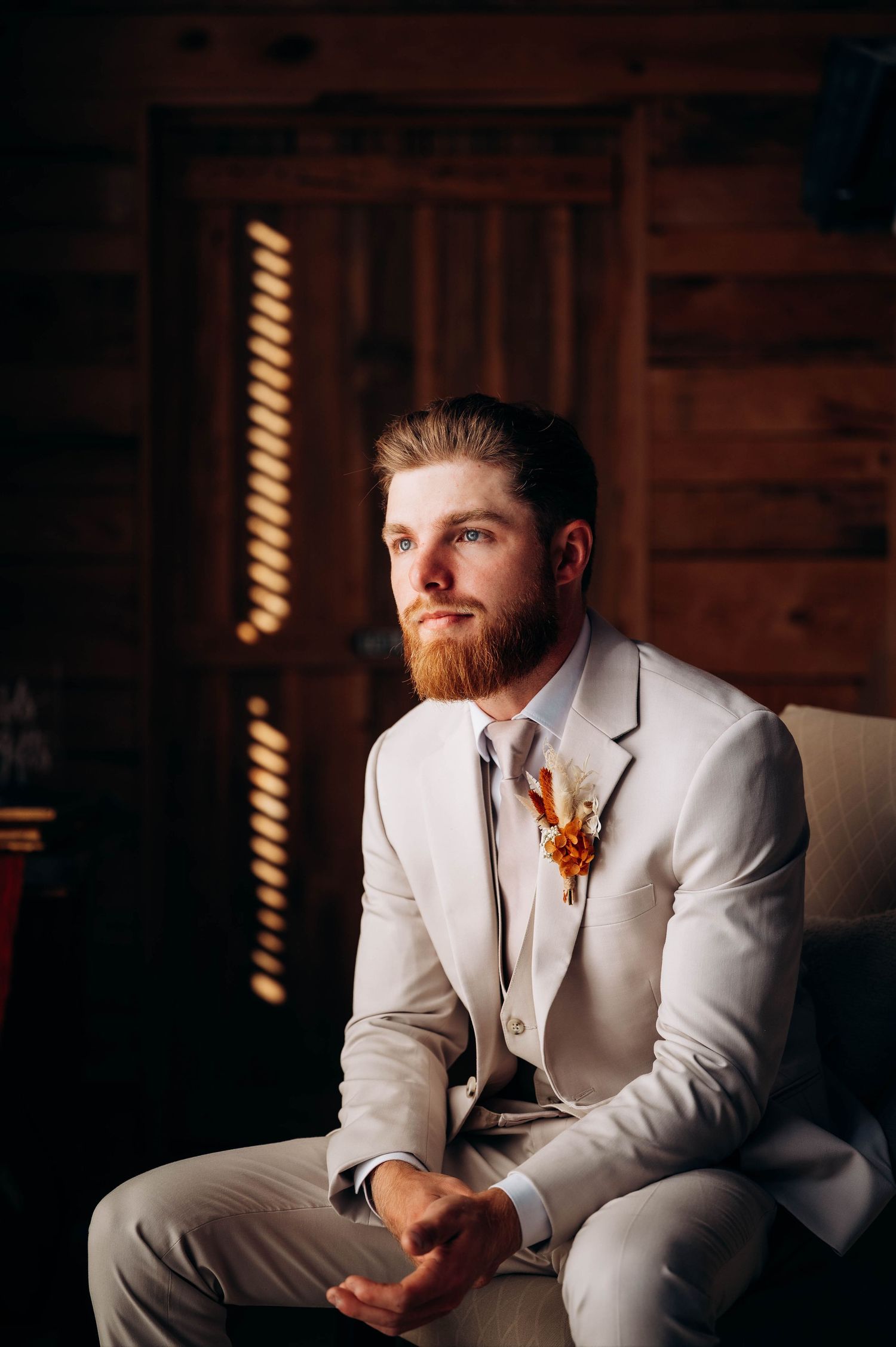 A groom in a white suit jacket sits thoughtfully in moody lighting during his wedding day.