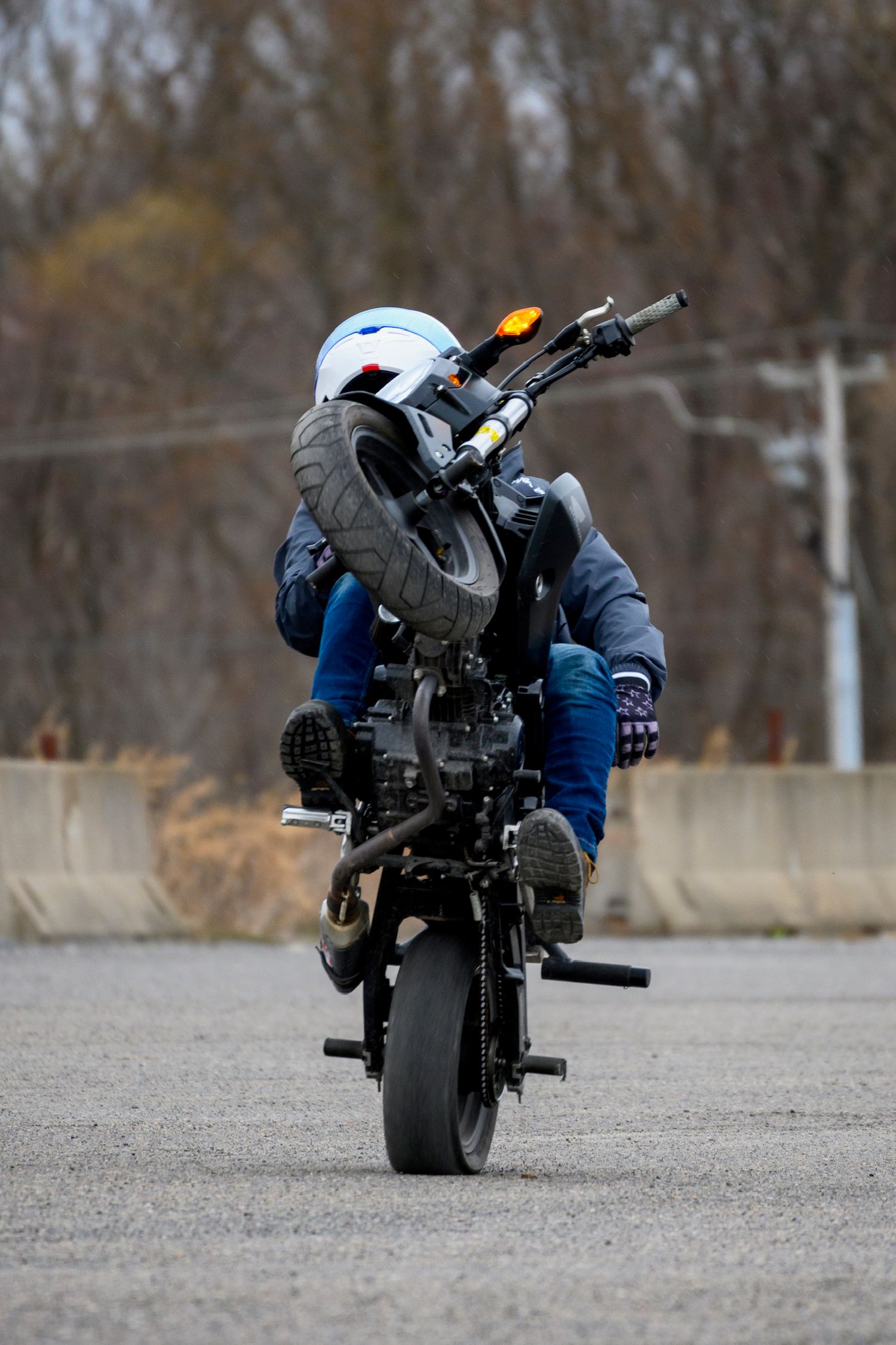 Motorcyclist performing a stoppie stunt on a black motorcycle in an empty parking lot wearing helmet and jeans.