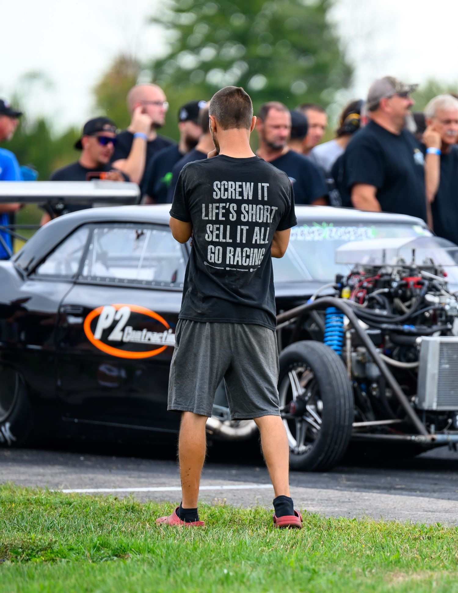 Spectators and racing enthusiasts gather around custom cars at an outdoor motorsport event.
